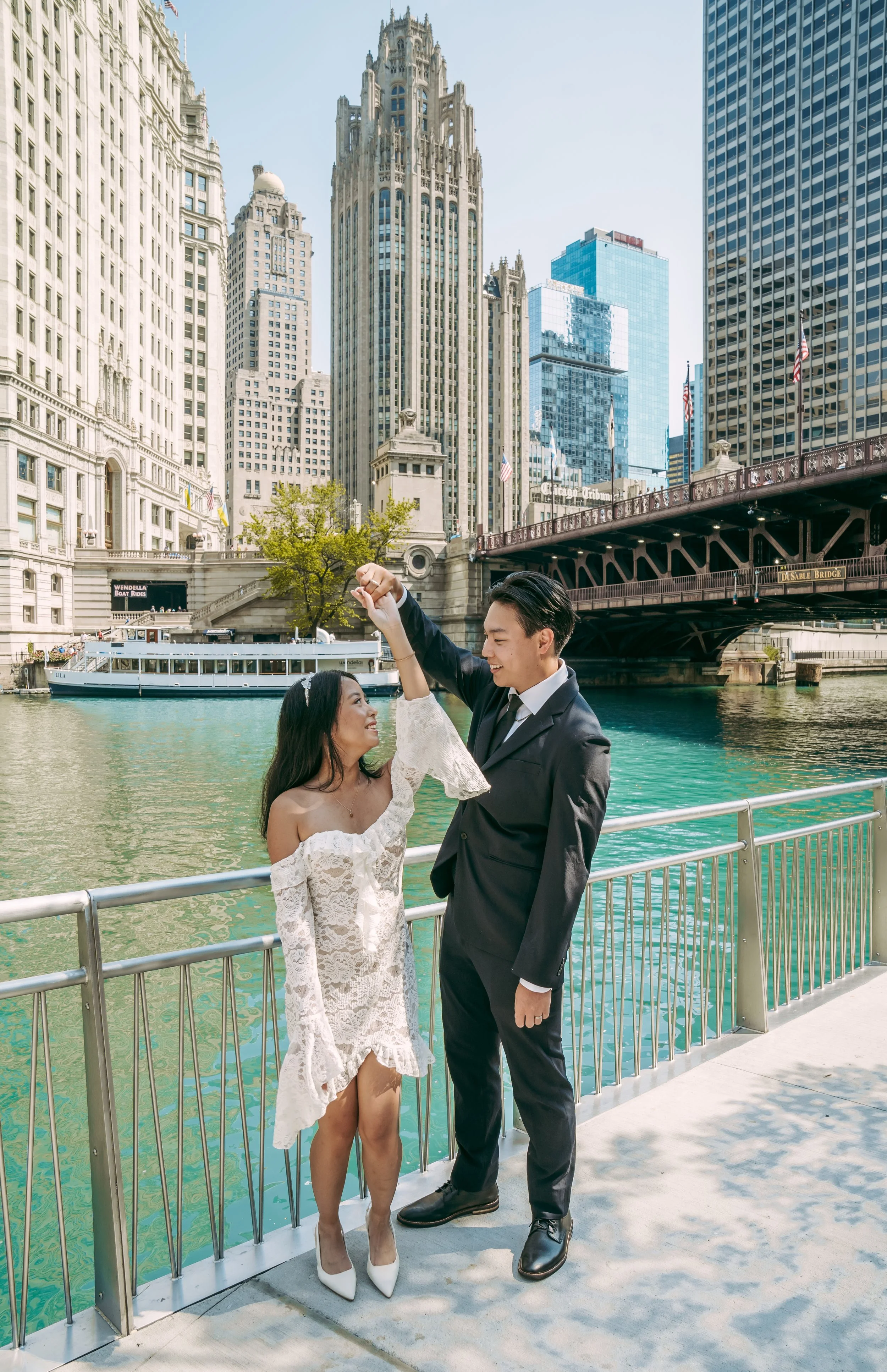A couple dressed in formal attire dancing by a river in a city with tall buildings and a bridge.