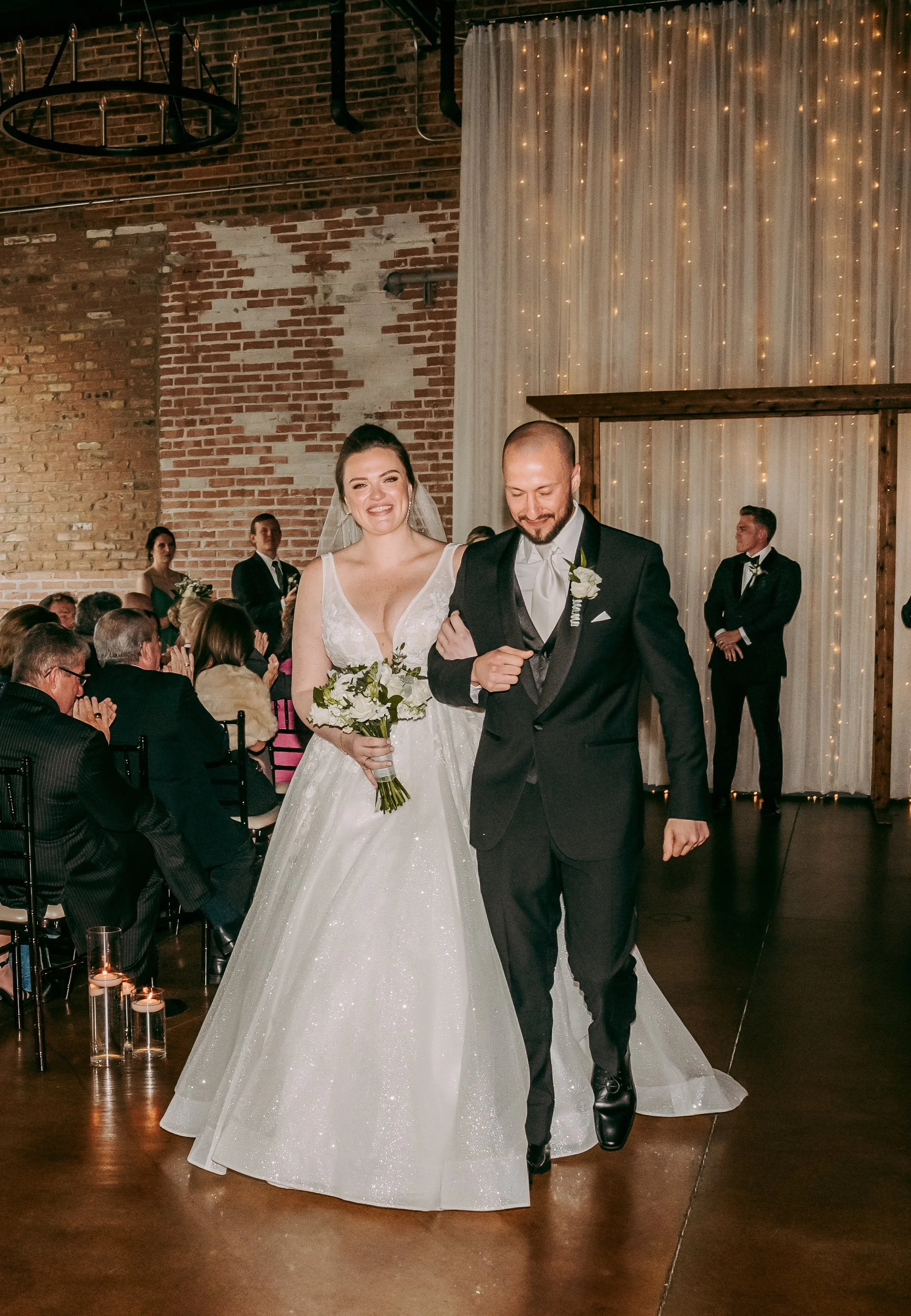 A bride and groom walking down the aisle at their wedding reception, smiling and holding hands, with guests clapping and a decorated backdrop behind them.