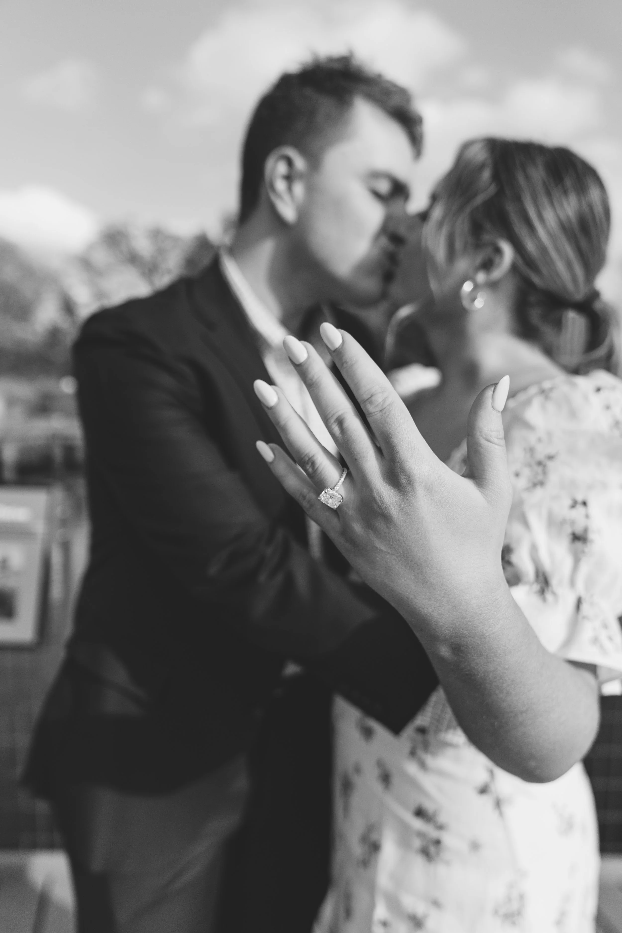 A woman showing an engagement ring on her finger and a couple kissing in the background.