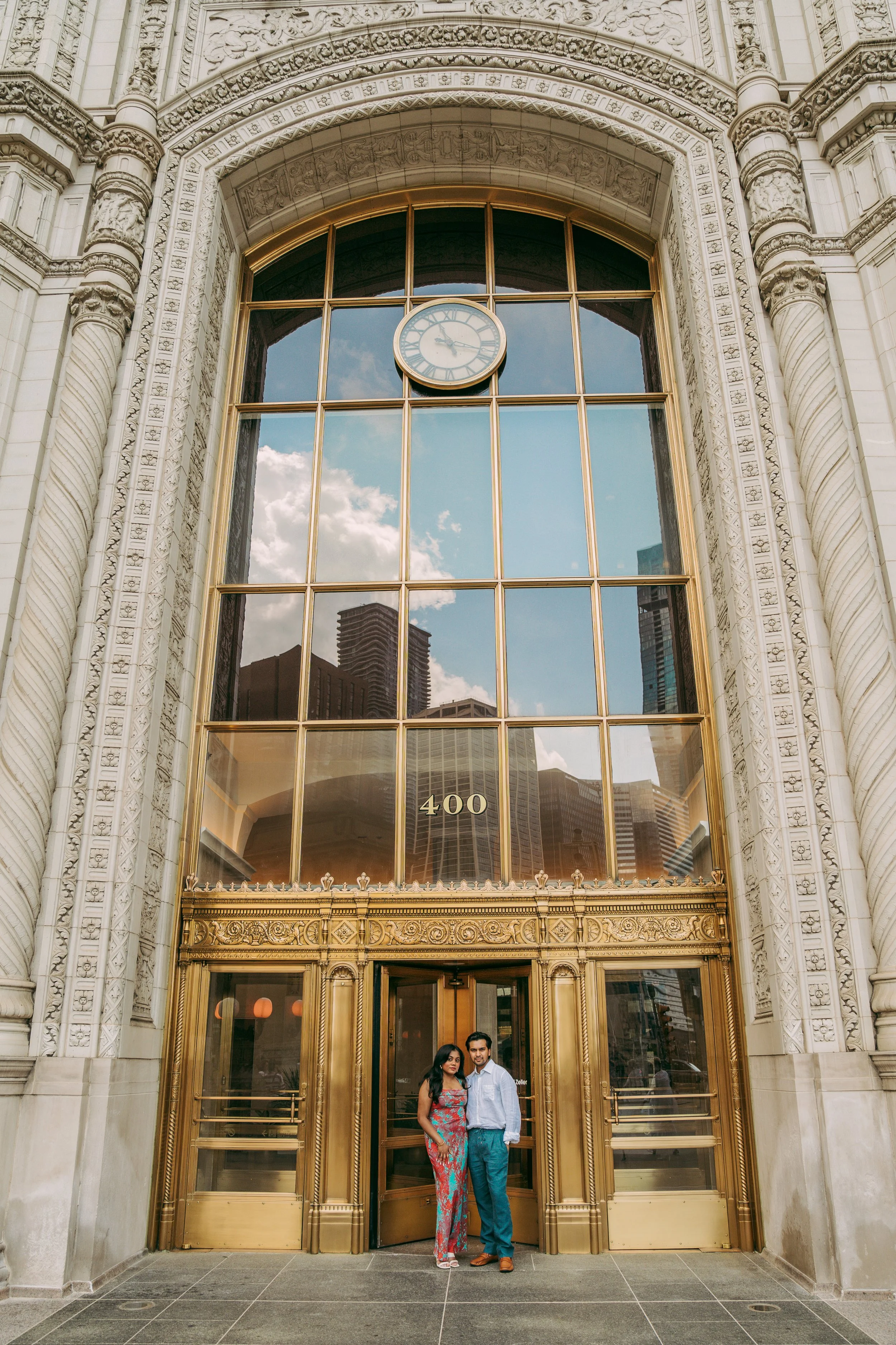 Two people standing in front of an ornate golden entrance of a building with large glass windows reflecting the city skyline and a clock showing nearly 2:13.