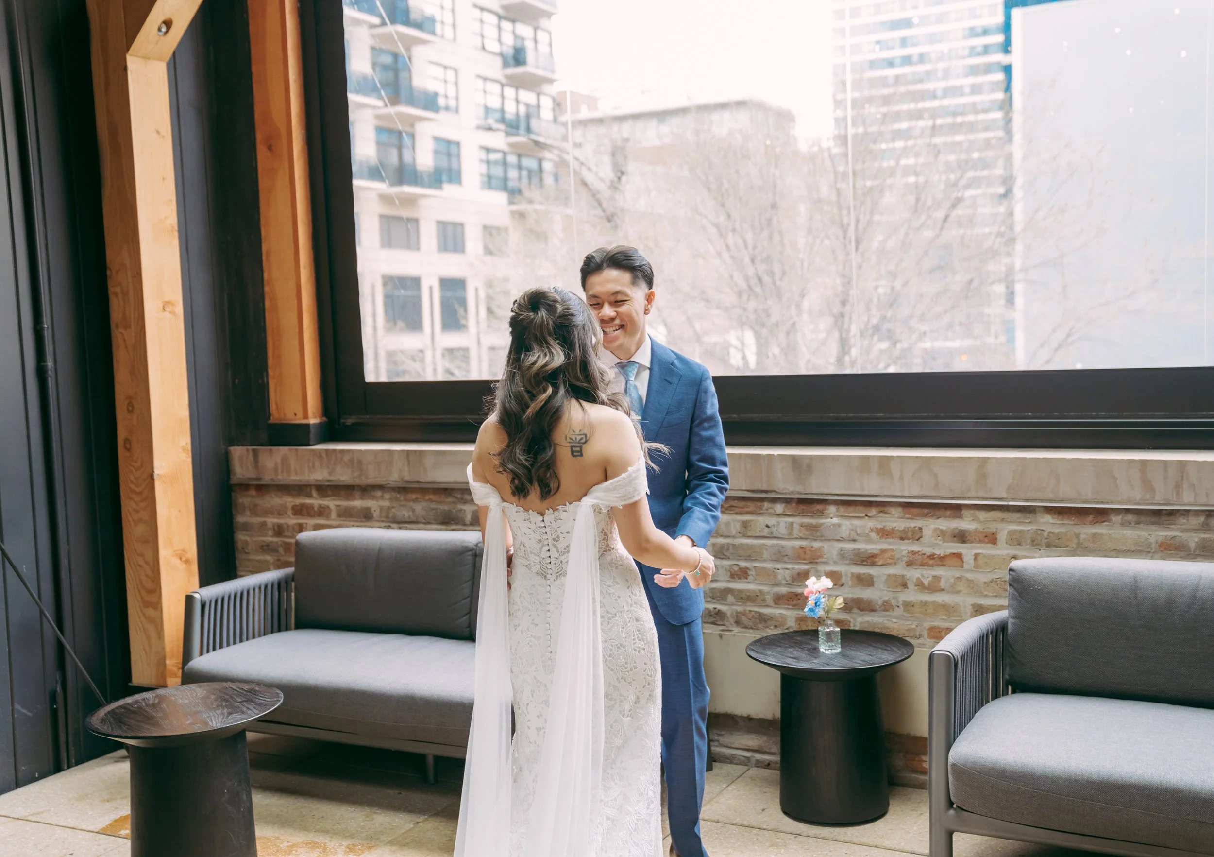 A couple, dressed in wedding attire, holding hands and smiling at each other in an indoor setting with large windows, brick wall, and modern furniture.