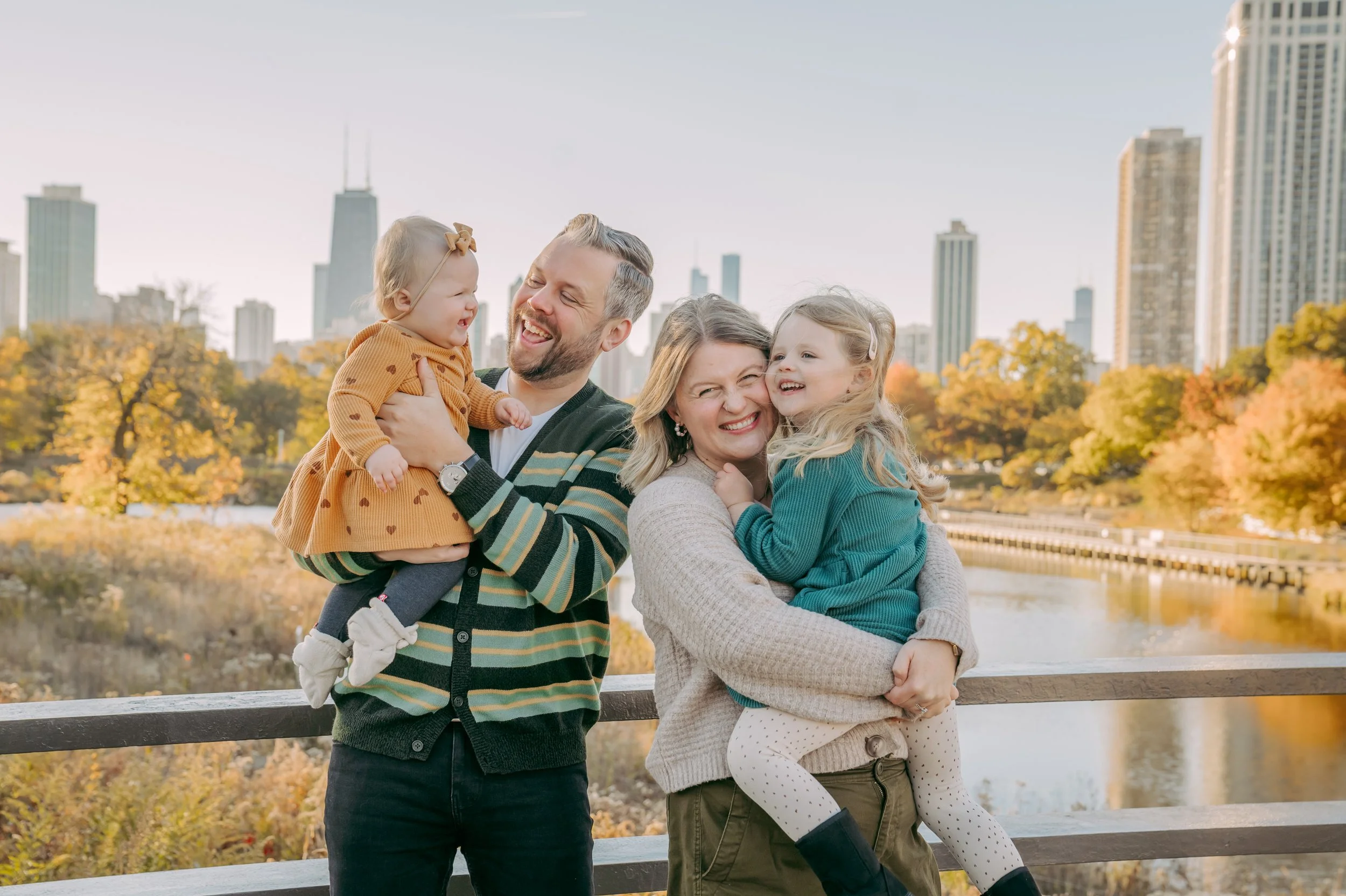 A happy family of four enjoying a sunny autumn day outdoors by a river, with city skyline and fall foliage in the background. The father holds a young girl, and the mother embraces a young girl, all smiling and laughing.