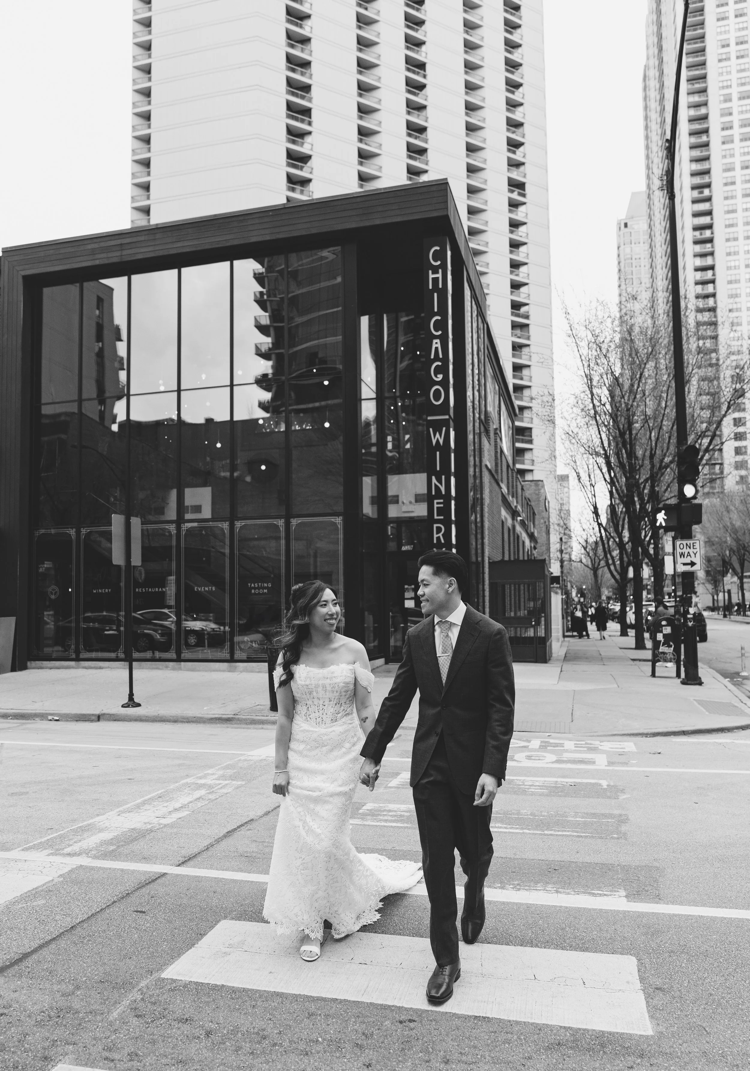 A black and white photo of a newlywed couple walking hand-in-hand across a city crosswalk in front of a building with a sign that says 'Chicago Winery'. The bride is wearing a strapless lace wedding dress, and the groom is in a suit and tie, both smi