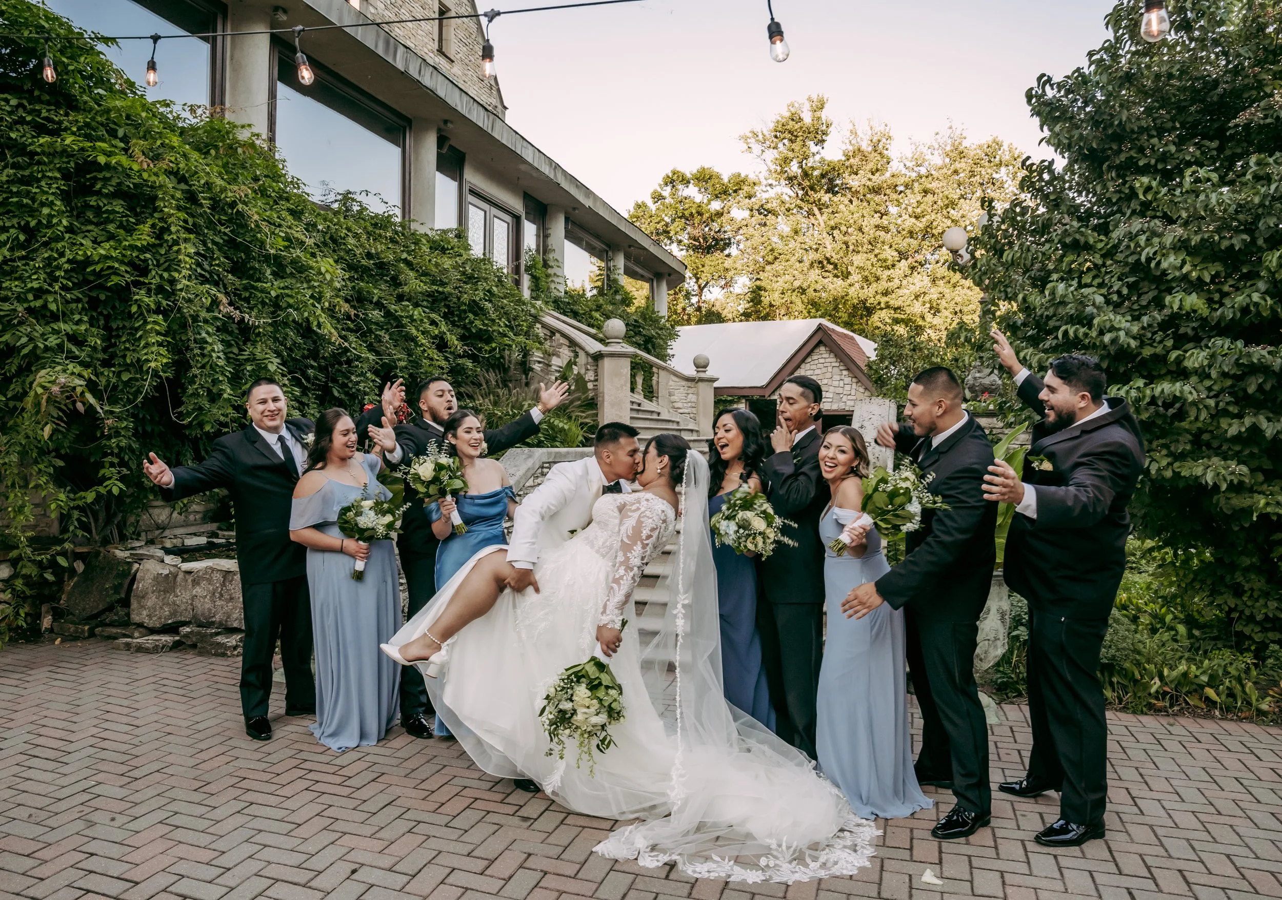 A wedding party of twelve people, including the bride and groom, celebrating outdoors on brick paving with greenery and a building in the background. The bride is being held by the groom, and everyone is smiling, cheering, and holding bouquets.