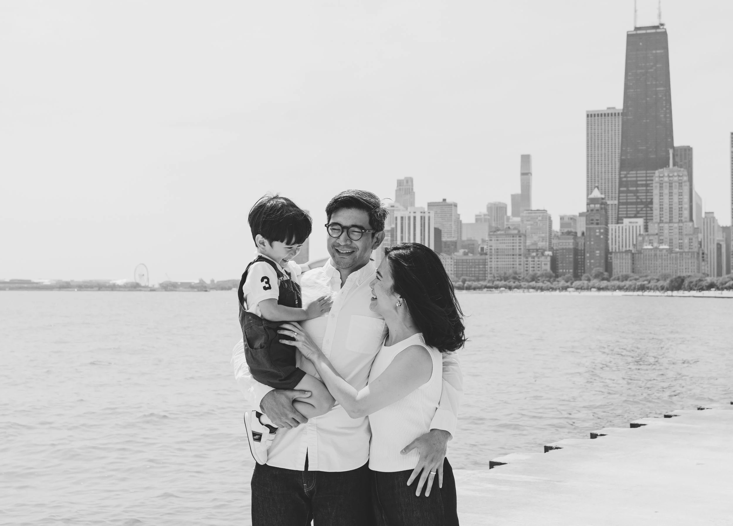 A happy family of three, a man, woman, and young boy, standing near a waterfront with a city skyline in the background. The man is holding the boy, and they are all smiling and looking at each other.