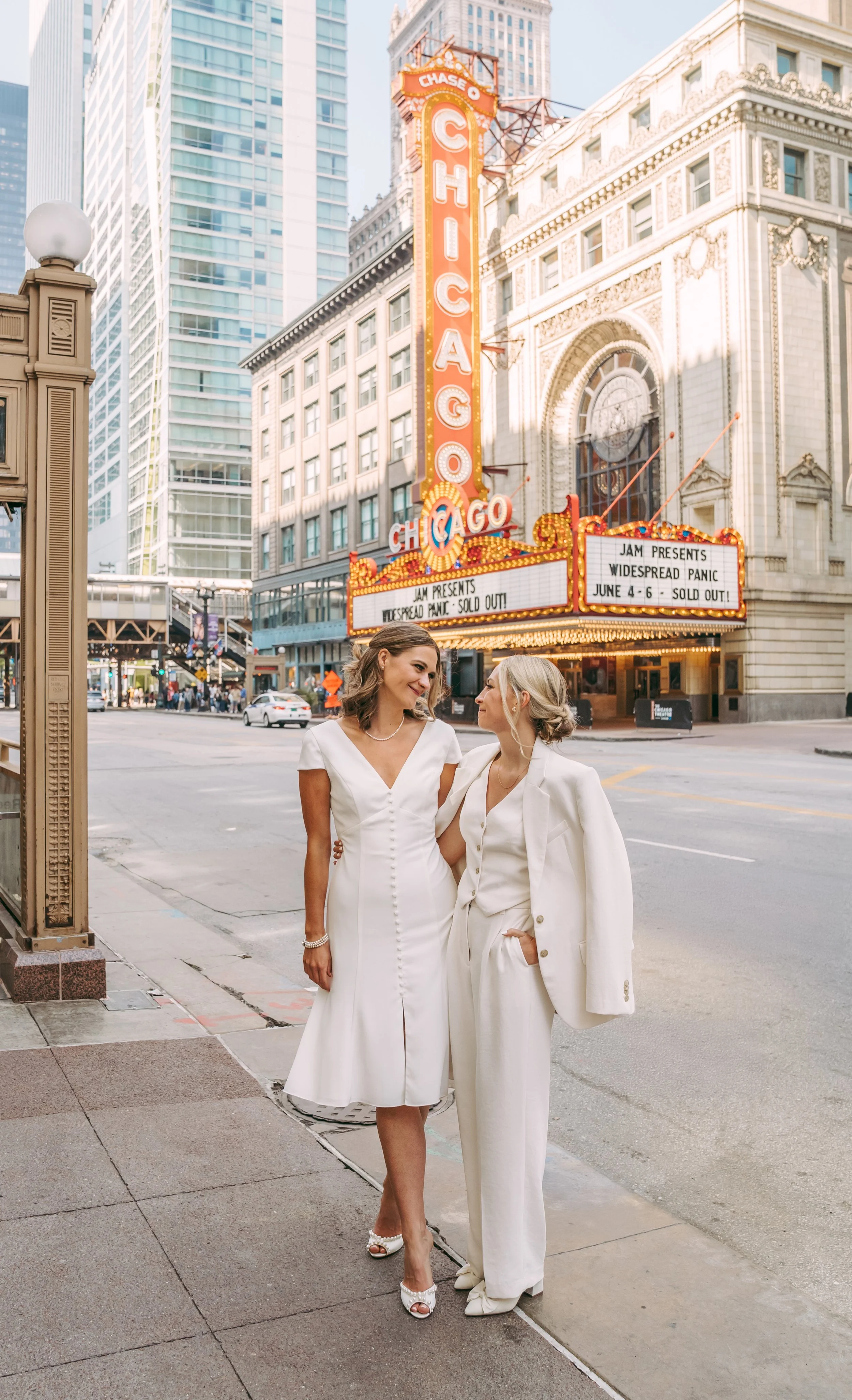 Two women dressed in white wedding attire standing on a city sidewalk in front of the Chicago Theatre marquee, looking at each other.