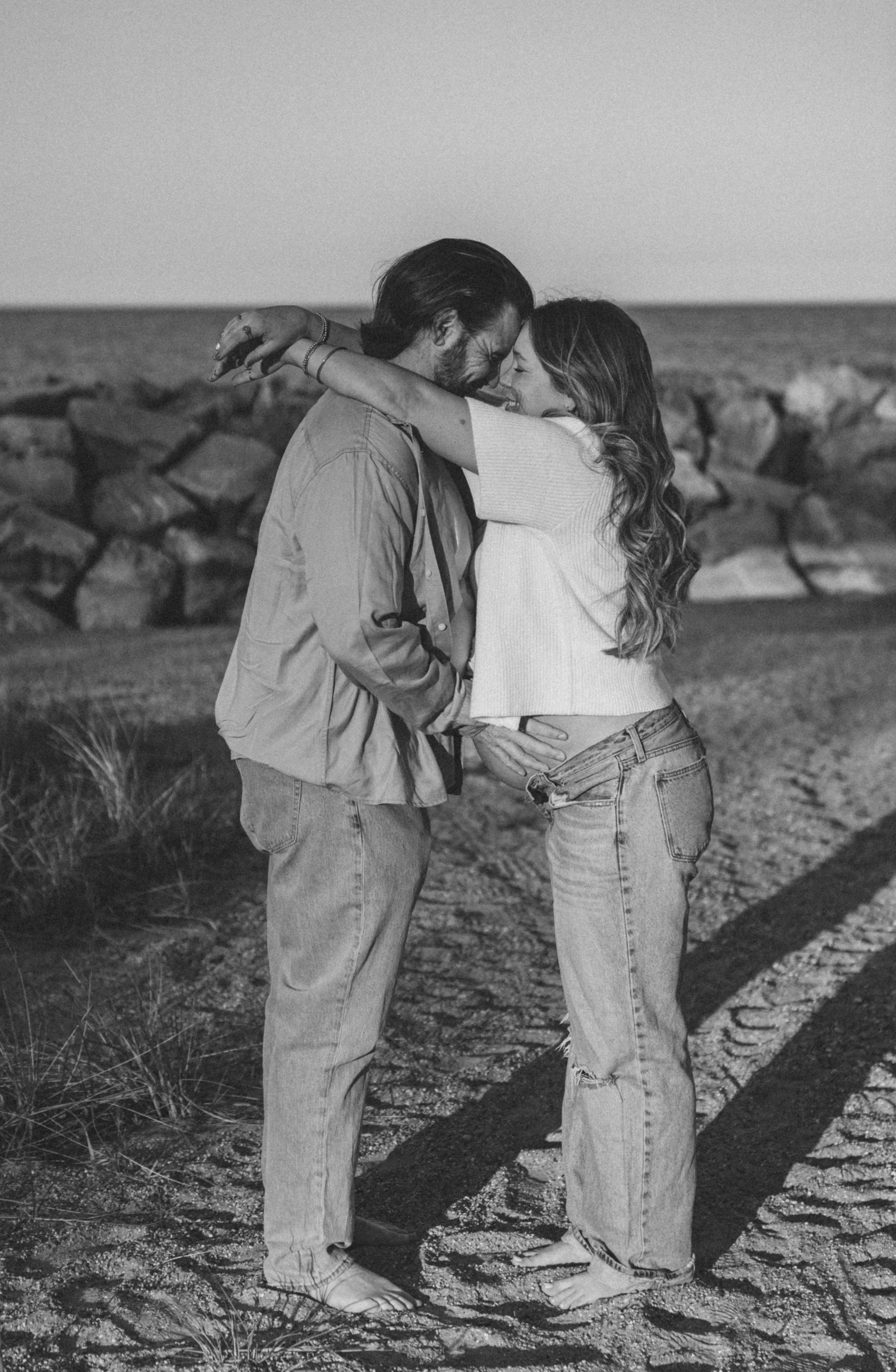 A black-and-white photo of a couple embracing at the beach, with rocks in the background and both individuals barefoot.