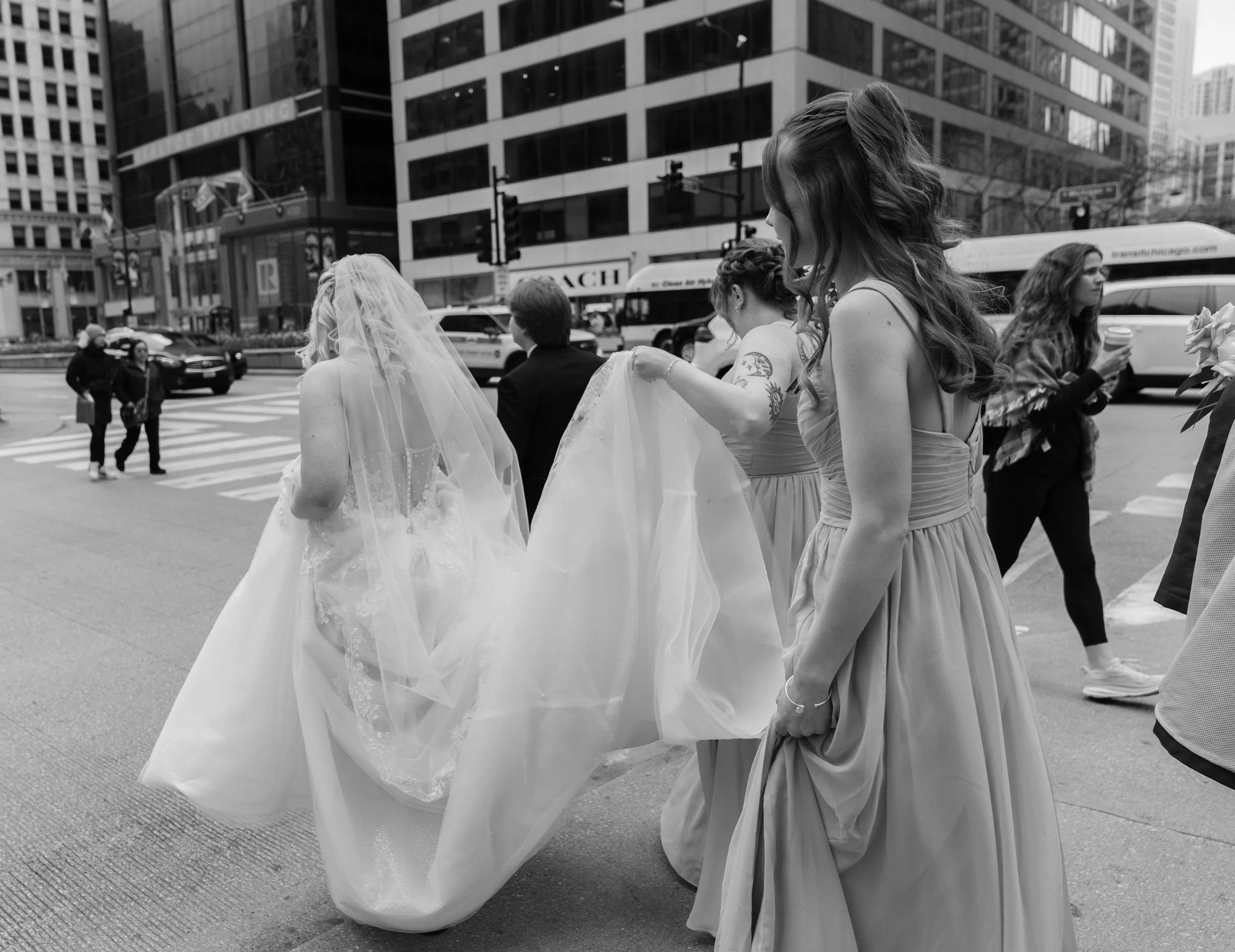 Group of women dressed in wedding and formal attire crossing a city street.