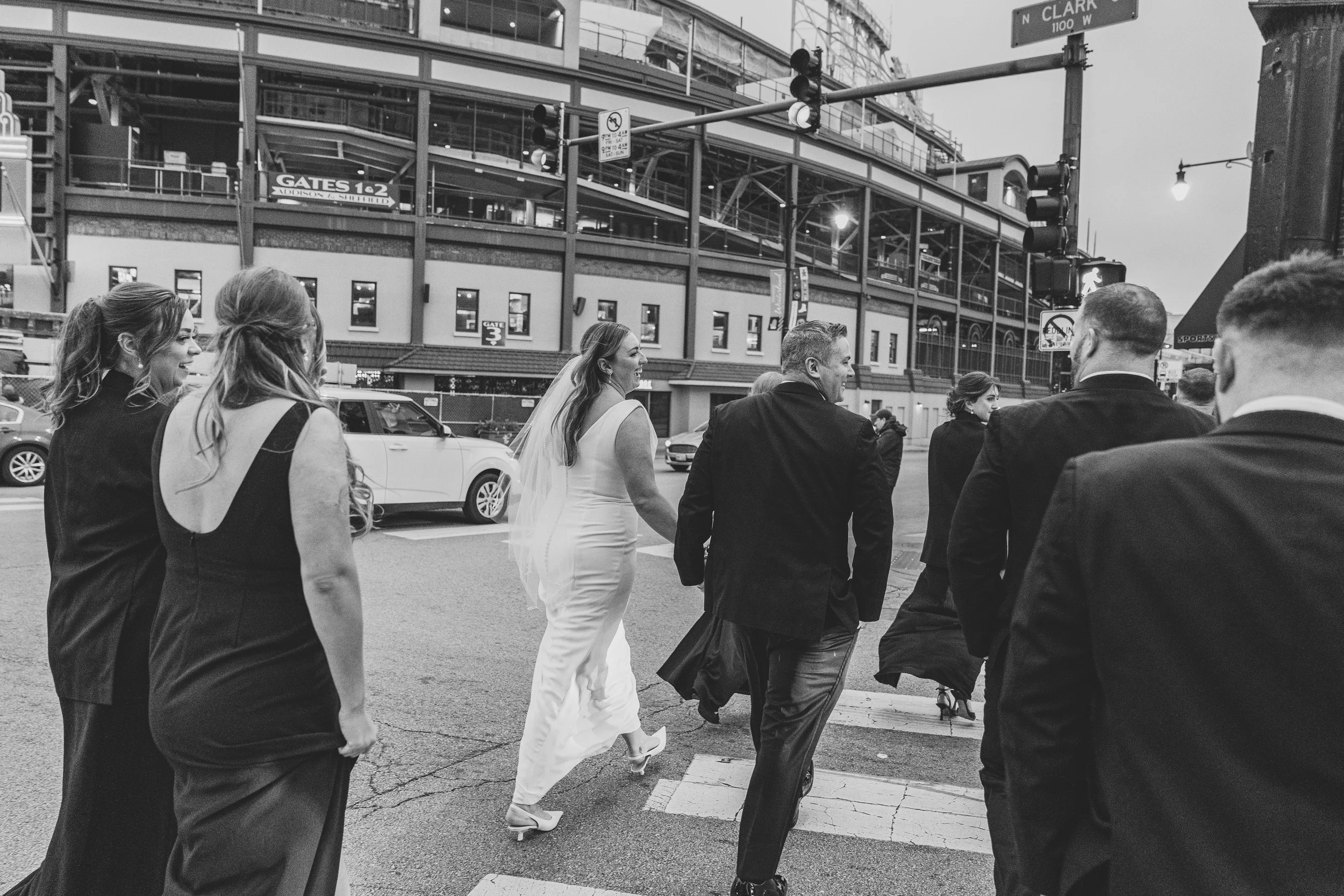 Black and white photo of a bride and groom walking across a city street with a group of people, possibly wedding guests, during the evening.