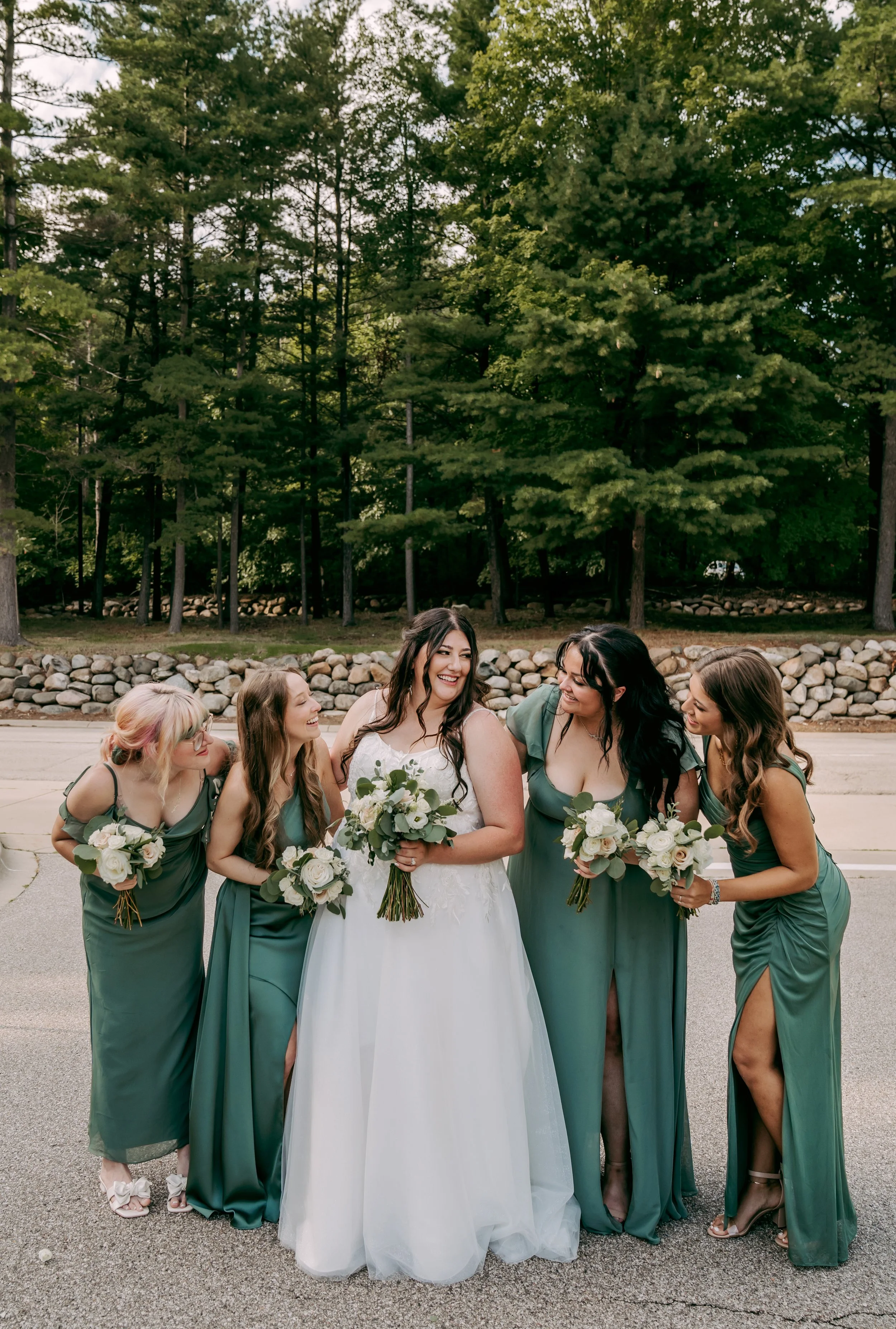 Group of five women, including a bride in a white gown and four bridesmaids in matching teal dresses, standing outdoors on a paved road with a stone wall and tall green trees in the background. They are smiling and holding bouquets of white flowers a