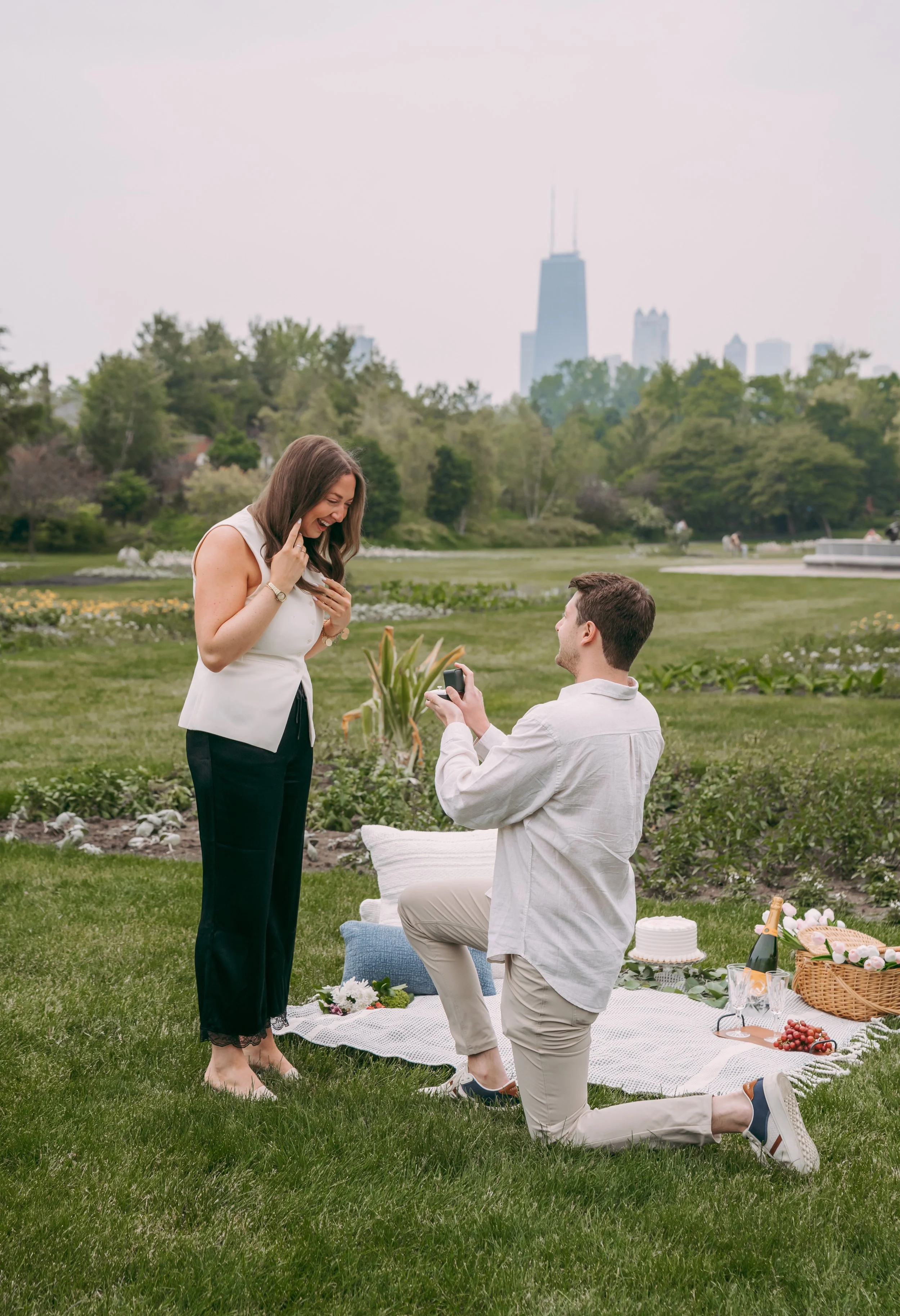 A man proposing marriage to a woman during a picnic in a park with city buildings in the background.