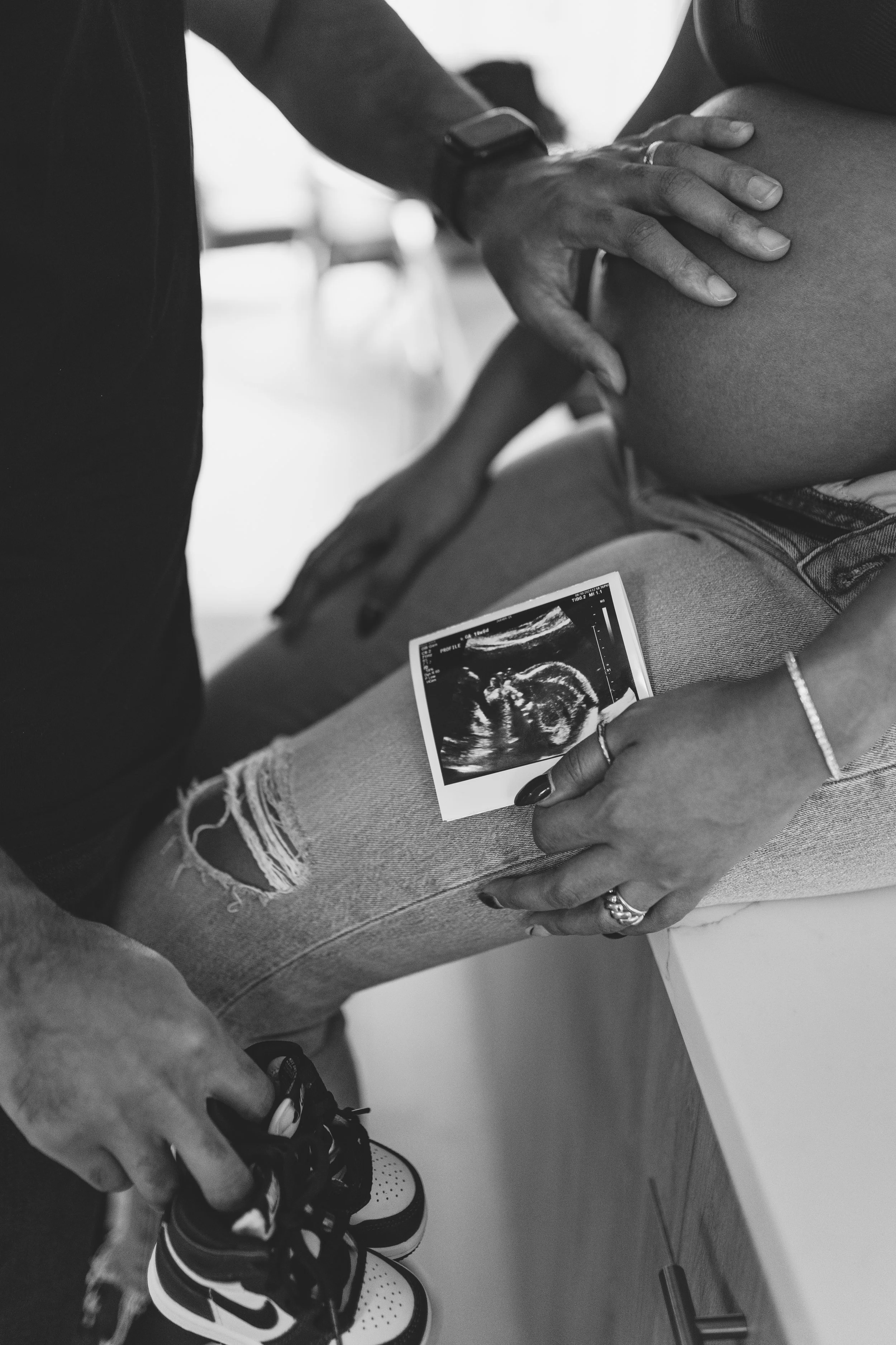 A pregnant woman holding an ultrasound photo and a man touching her belly, seated on a table, in black and white.