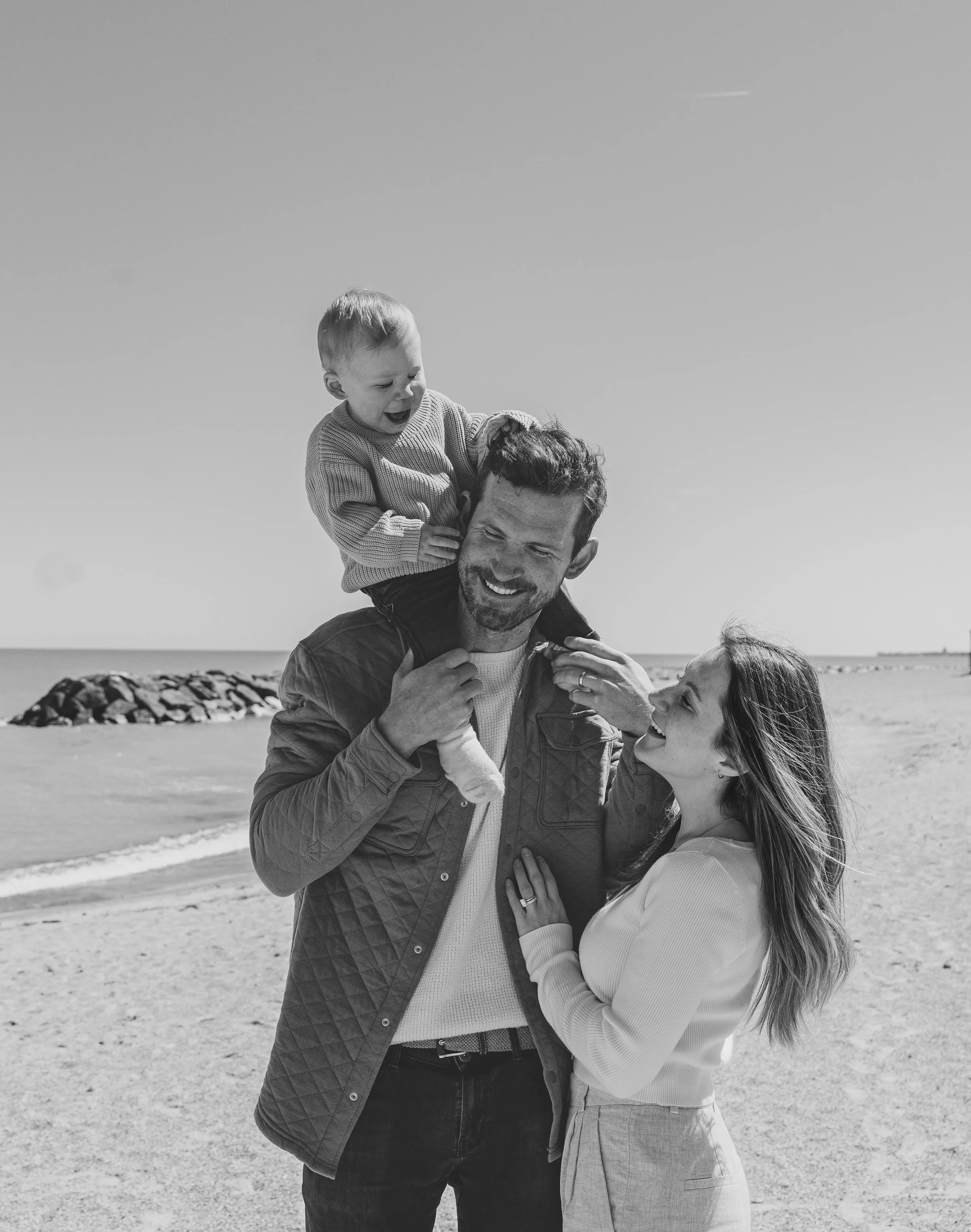 Black and white photo of a family at the beach: father carrying a toddler on his shoulders, mother smiling at him, with the ocean and rocks in the background.