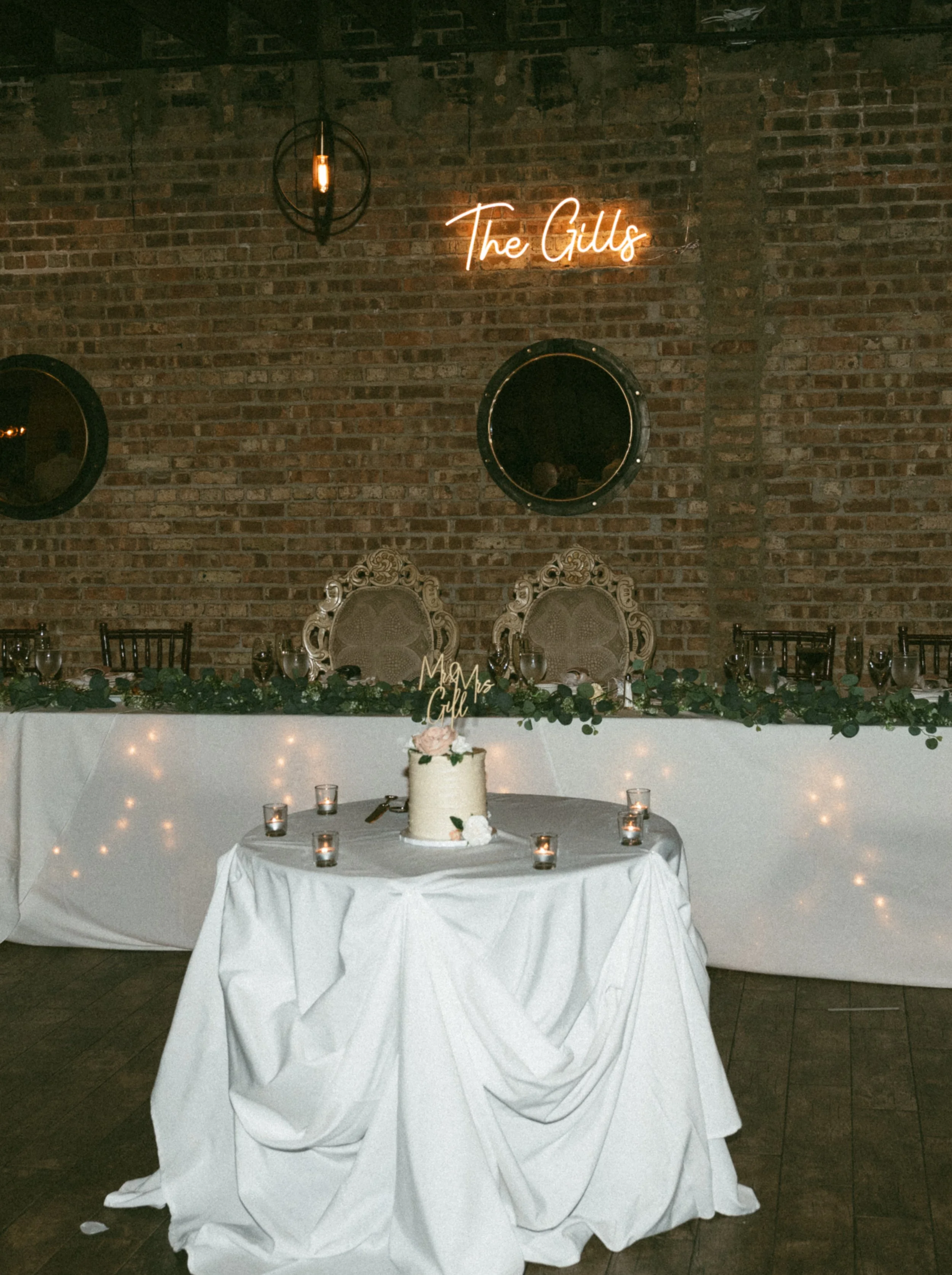 Wedding reception table with a cake, candles, and flowers against an exposed brick wall with a neon sign that reads 'The Gill's'.