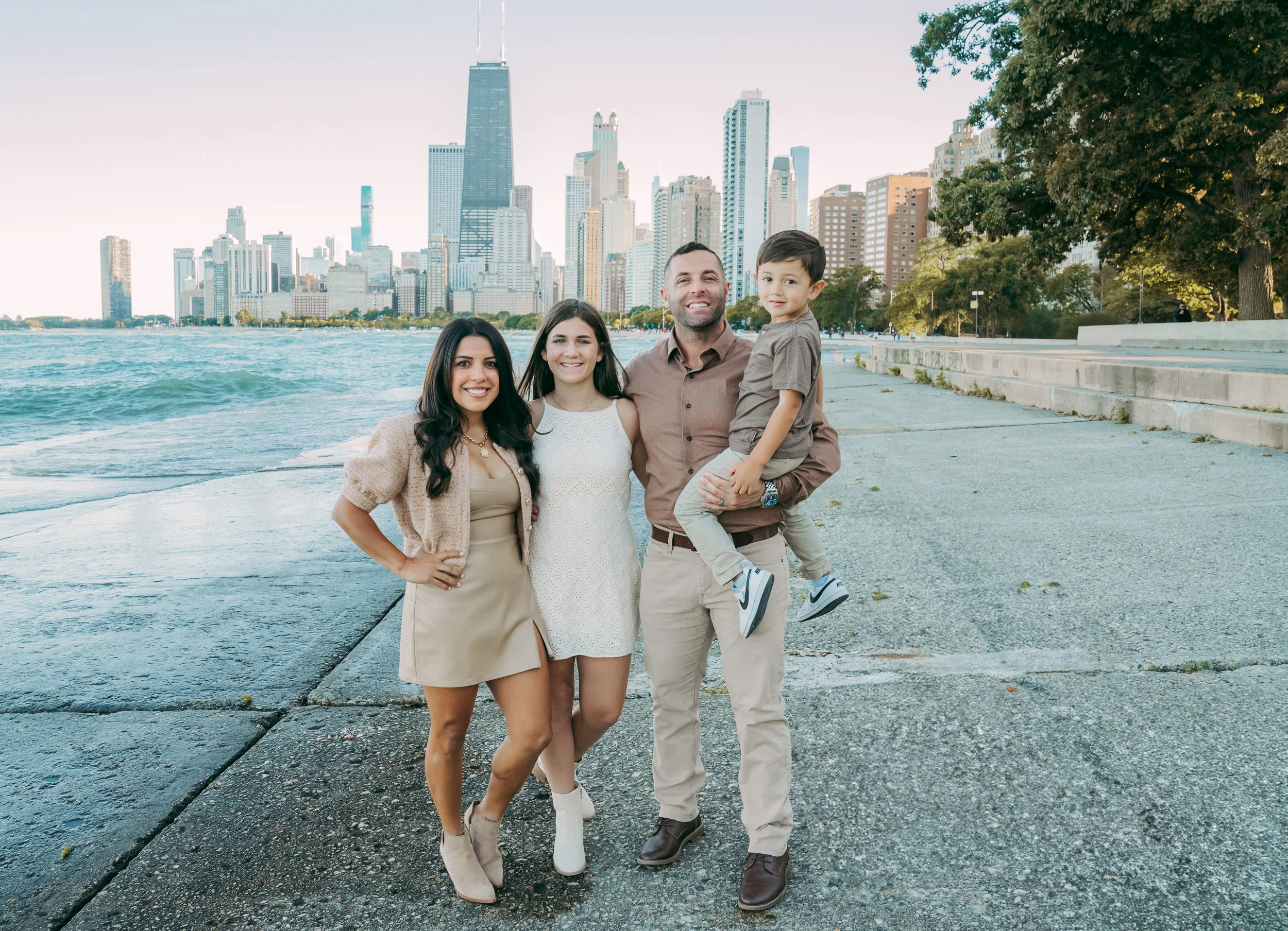 Family of four standing by Lake Michigan with Chicago skyline in background, smiling, during daytime.