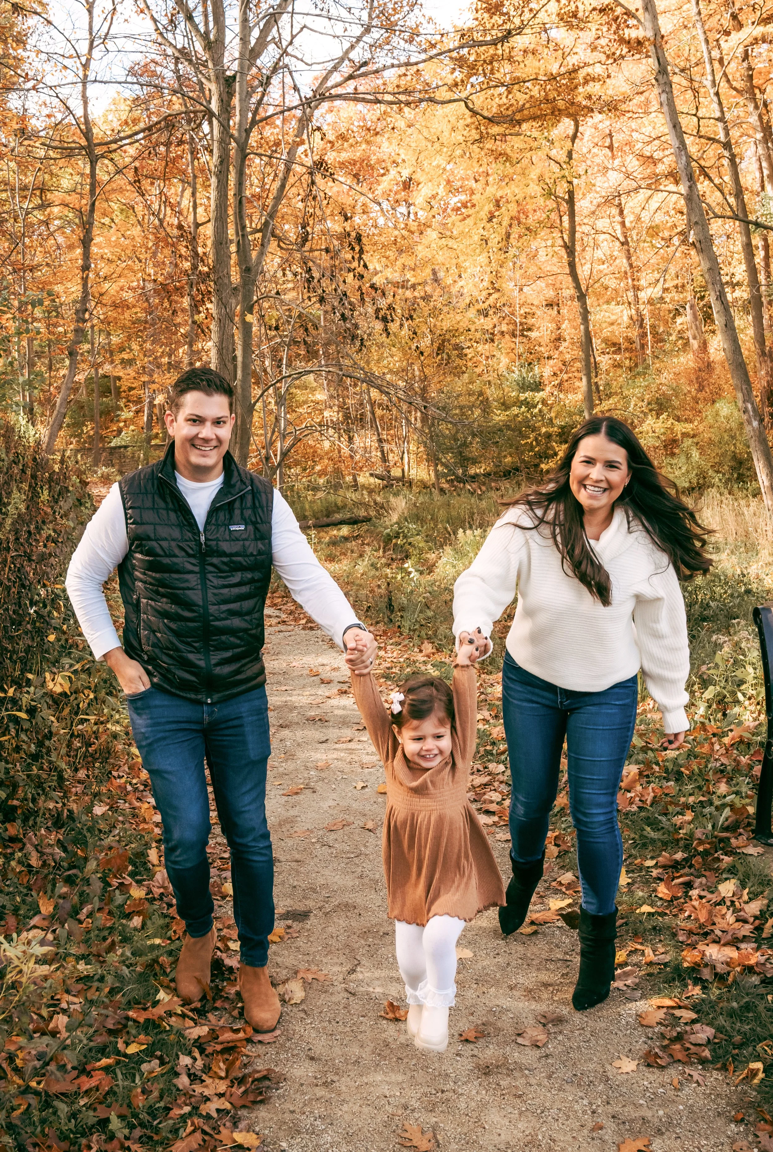 A family of three, a man, a woman, and a young girl, walking hand-in-hand outdoors on a fall day near trees with orange leaves.