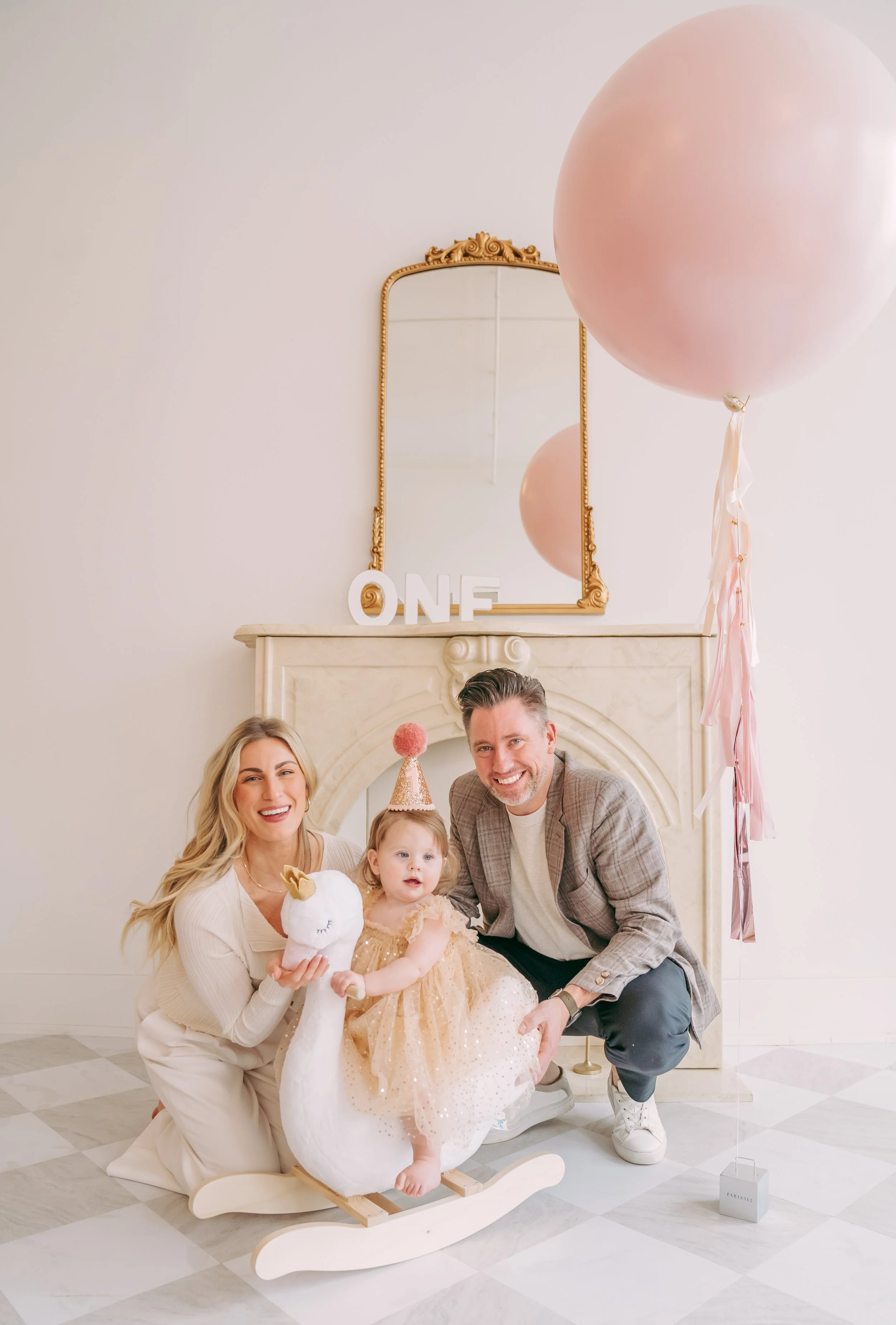 Family celebrating a first birthday with a plush unicorn toy, pink balloons, and decorations, smiling in front of a fireplace.