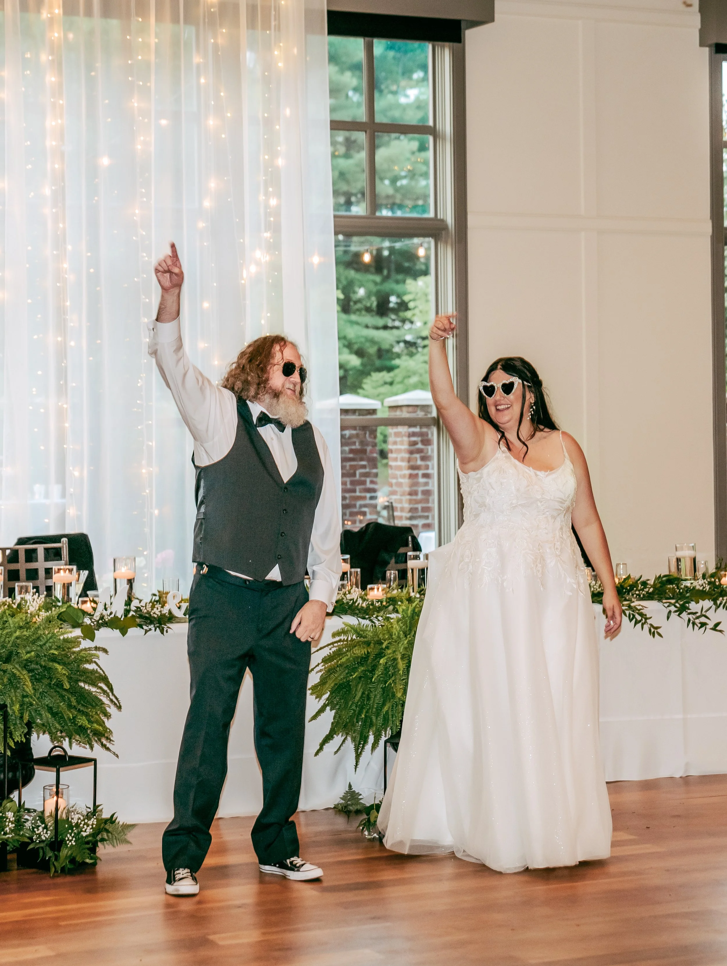 A man and woman, dressed in wedding attire, dancing and celebrating at a wedding reception with decorated tables and string lights in the background.