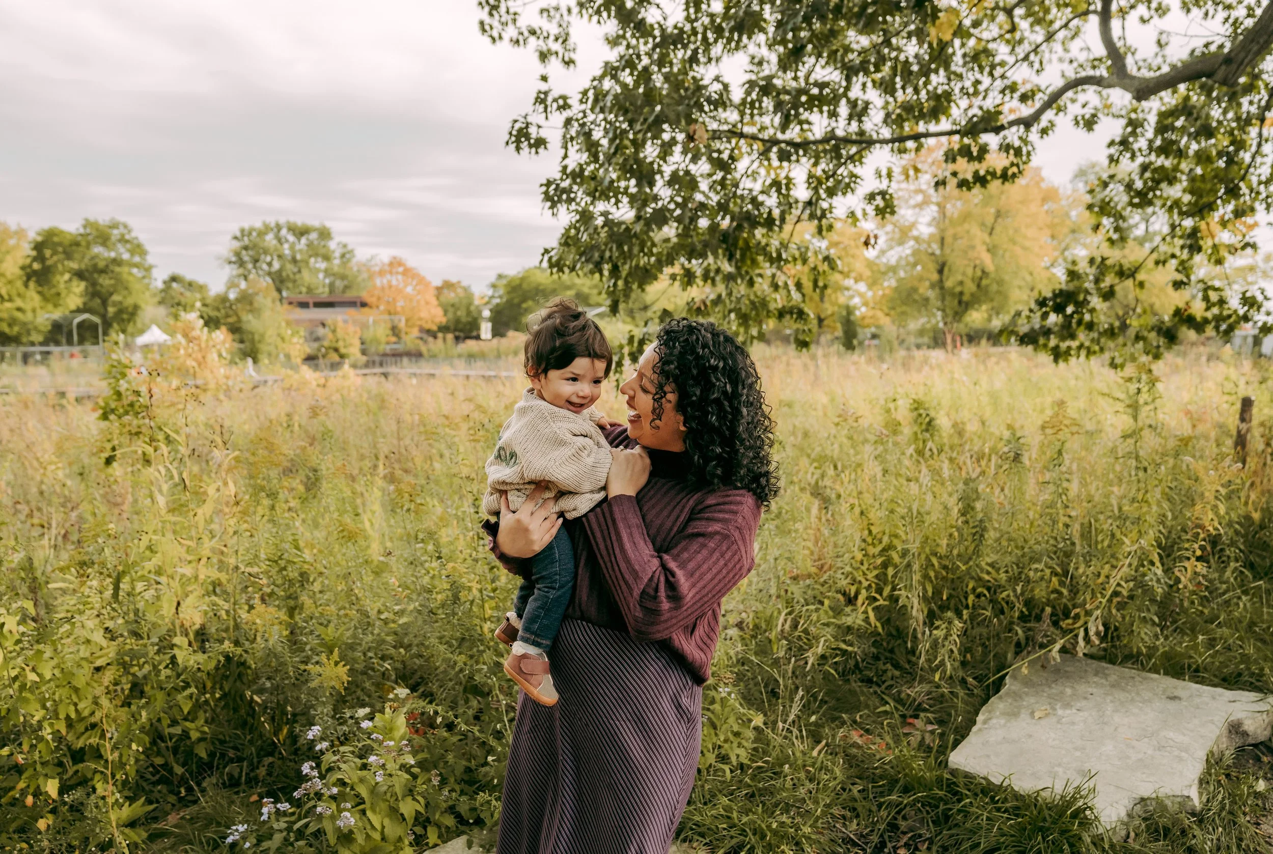 A woman holding a young child in a park with autumn foliage and cloudy sky.