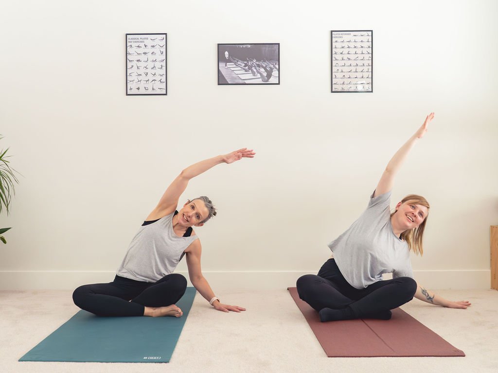 Two women practicing yoga indoors, sitting on yoga mats and performing side stretches while smiling.