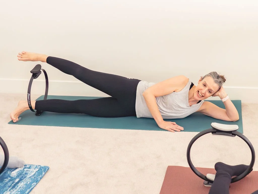 Older woman lying on her side on a yoga mat with Pilates rings around her ankles, engaging in a fitness activity.