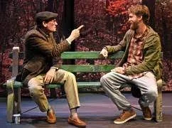 Two men sitting on a park bench having a conversation, surrounded by trees with red leaves, in a live theater production.