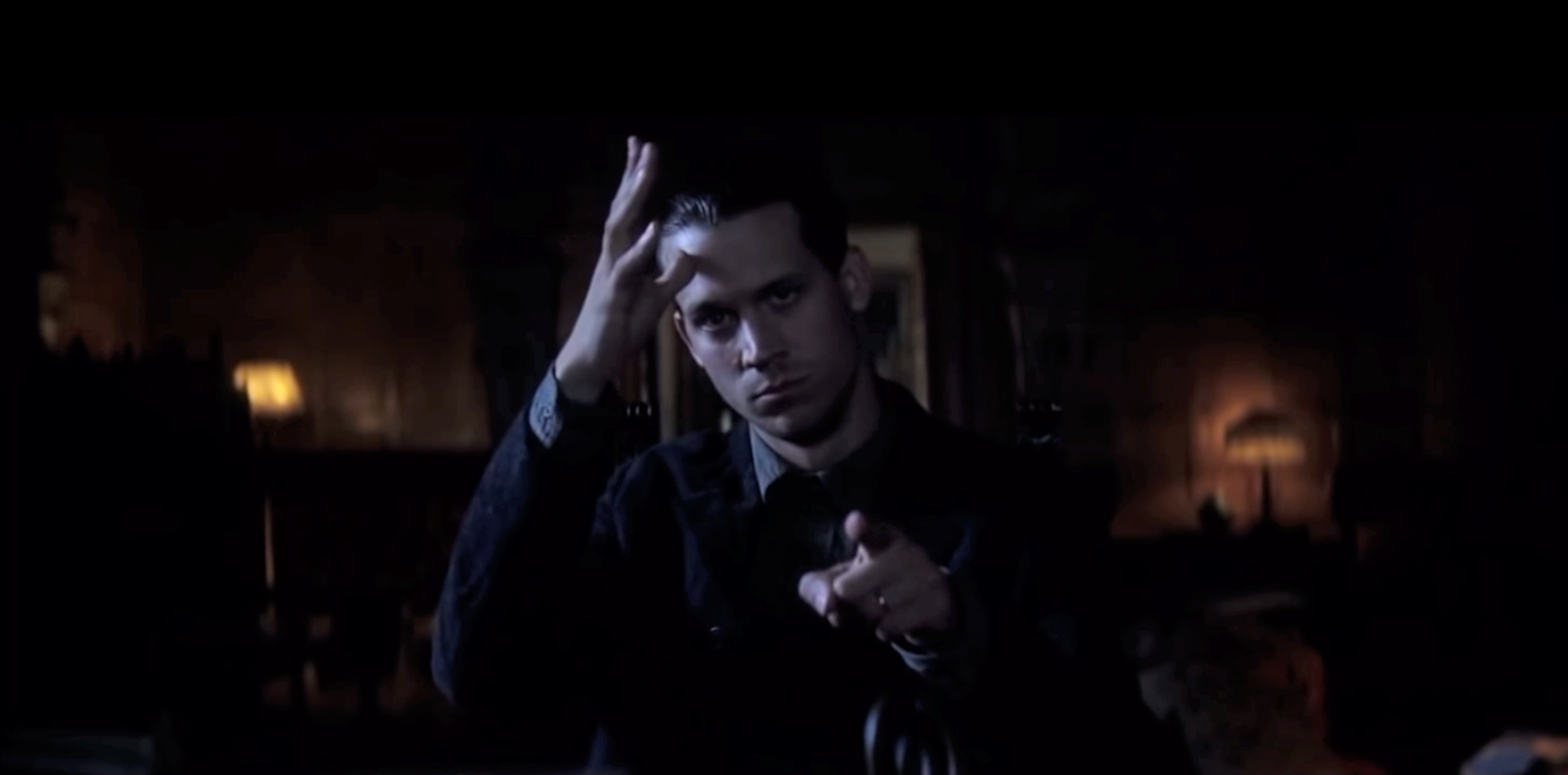 Russell Harvard (in character) wearing a dark jacket and shirt, pointing his hand upwards, with a serious expression, in a dimly lit room with warm lighting and lamps in the background.