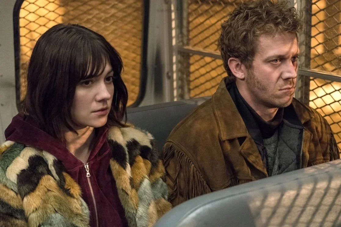 A woman with dark hair and a man with curly hair sit side by side on a bus or train with a metal grid wall behind them, looking serious or upset.