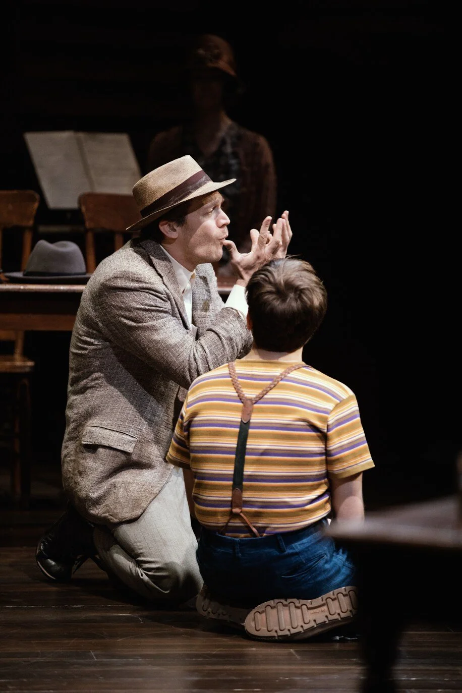 Russell Harvard (in character) kneeling on the floor, wearing a brown hat and a tweed jacket, appears to be explaining something, in sign language, to a young boy with short hair, dressed in a striped t-shirt and suspenders.
