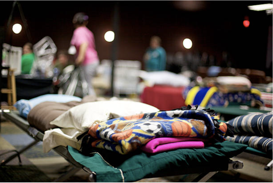Stacks of folded blankets and pillows on cots at a thrift store or donation center with blurred people in the background.