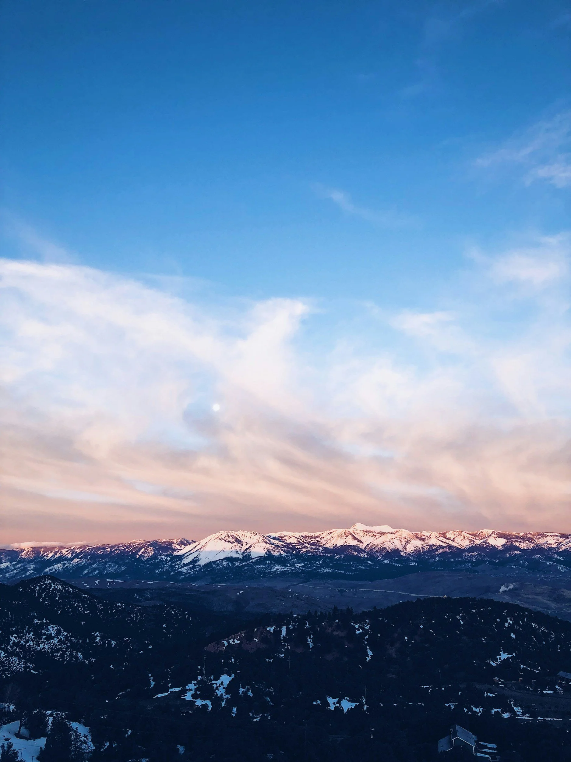 Snow-capped mountains under a partly cloudy sky during sunset with the moon visible.