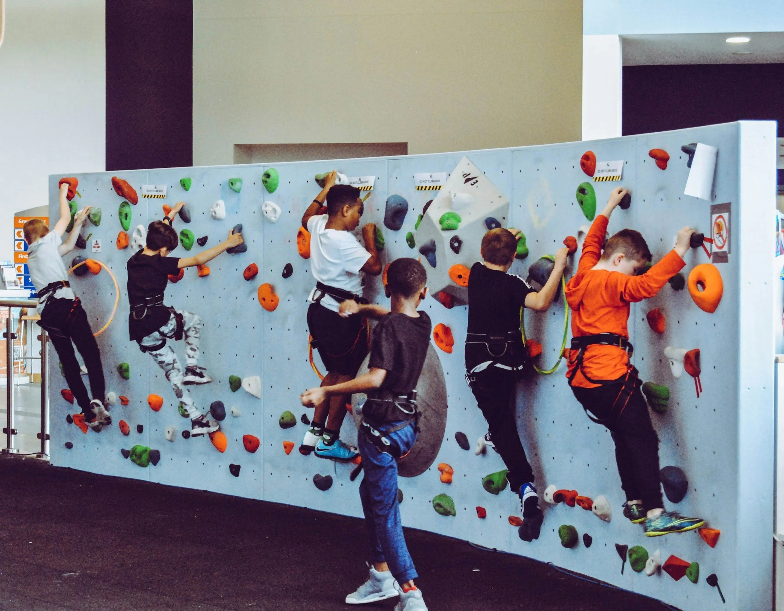 Six children of diverse ethnicities climbing an indoor rock climbing wall with colorful holds, in a recreational facility.