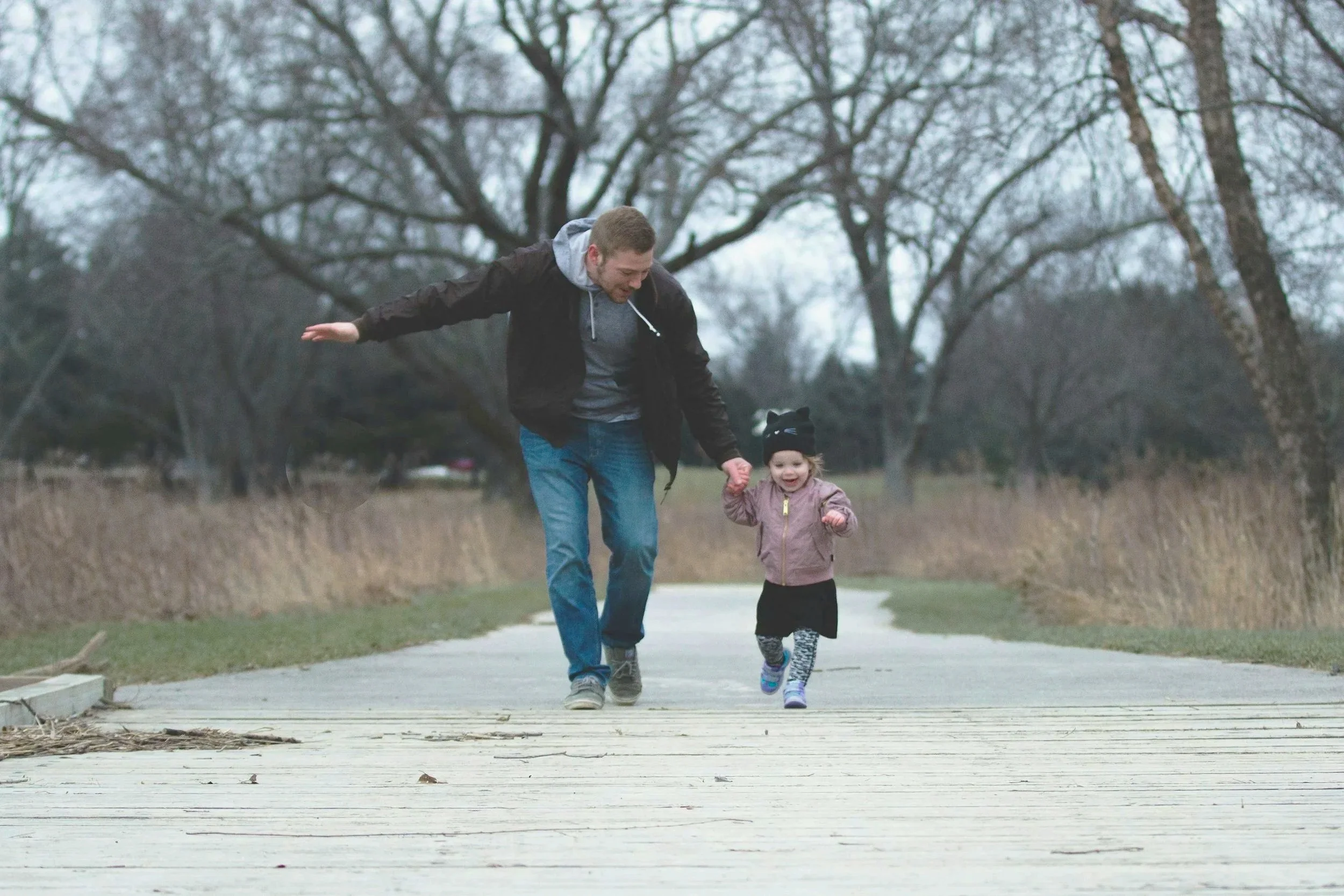 A man and a young girl walking hand-in-hand on a wooden path in a park during late fall or winter.