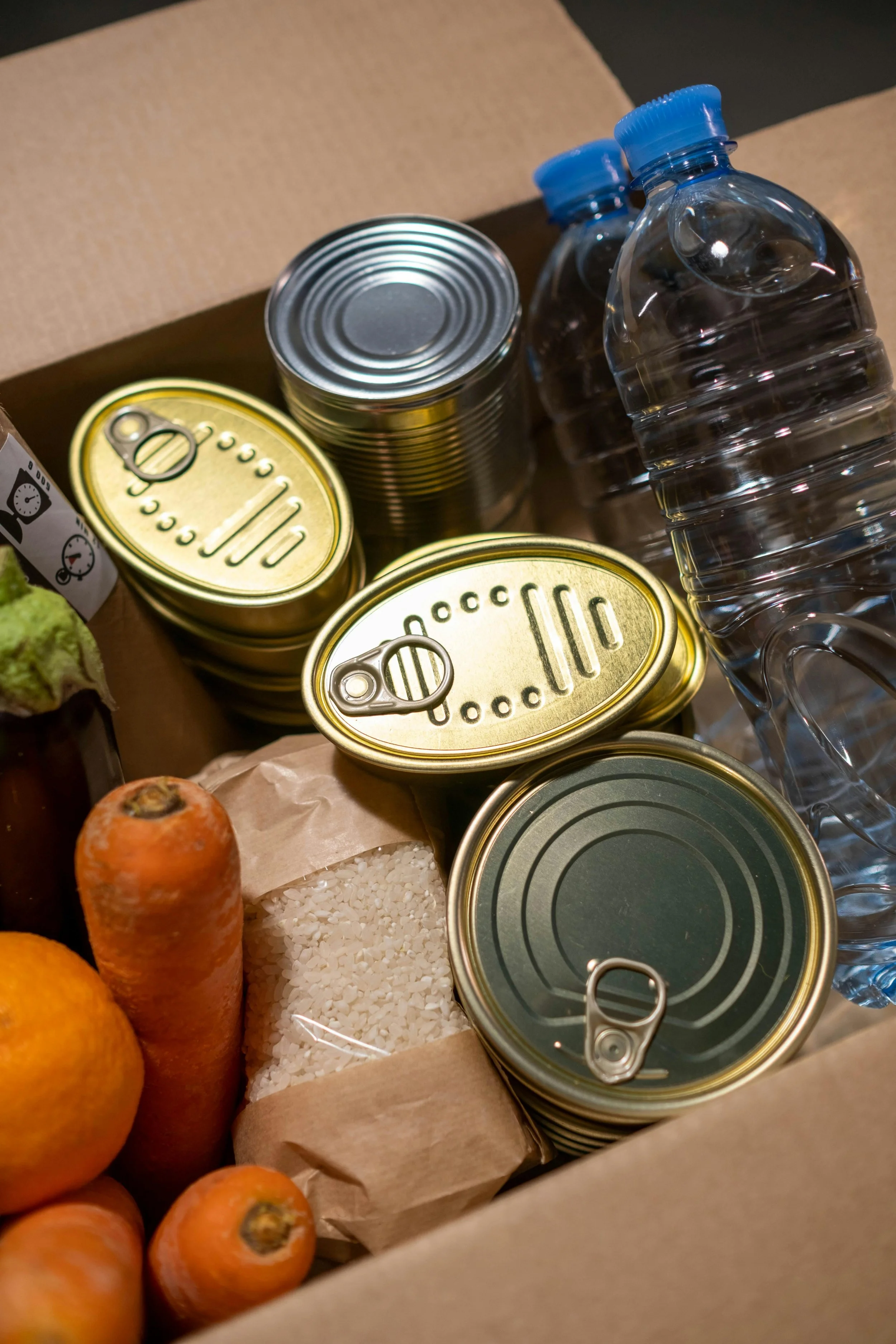 A cardboard box filled with canned vegetables, bottled water, fresh carrots, oranges, salt, and other grocery items.