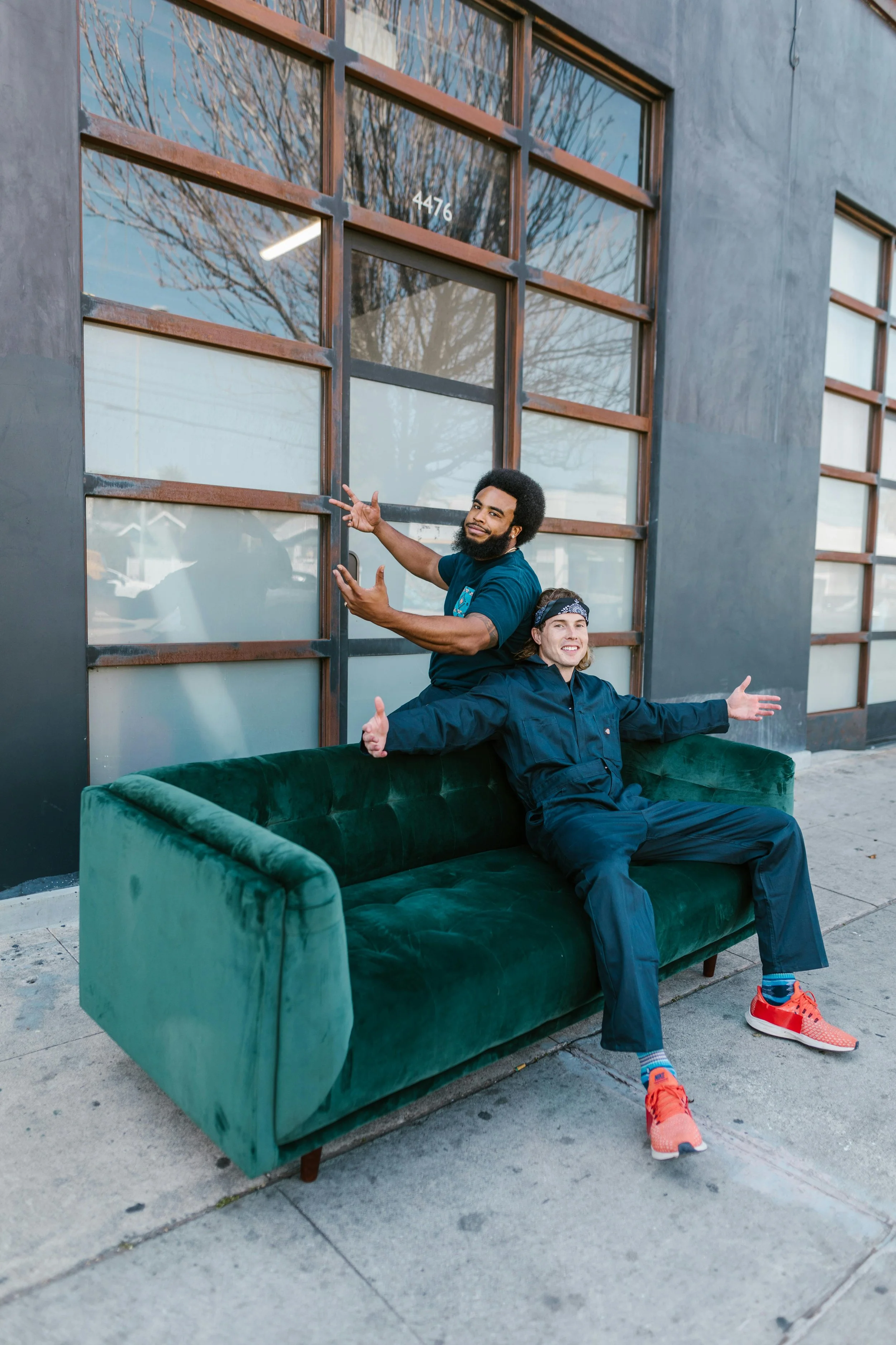 Two men are sitting on a green velvet couch outside in front of a building with large windows. One man is sitting on the couch with arms outstretched, and the other is standing behind, smiling and gesturing toward the camera.