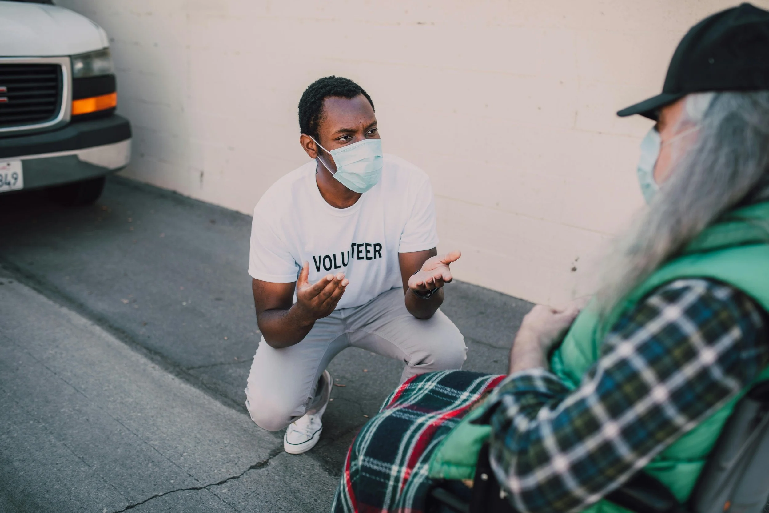 A young man wearing a white t-shirt with 'VOLUNTEER' written on it, kneeling and talking to an elderly person sitting in a wheelchair. Both are wearing face masks, and the scene takes place outdoors on pavement near a white wall.