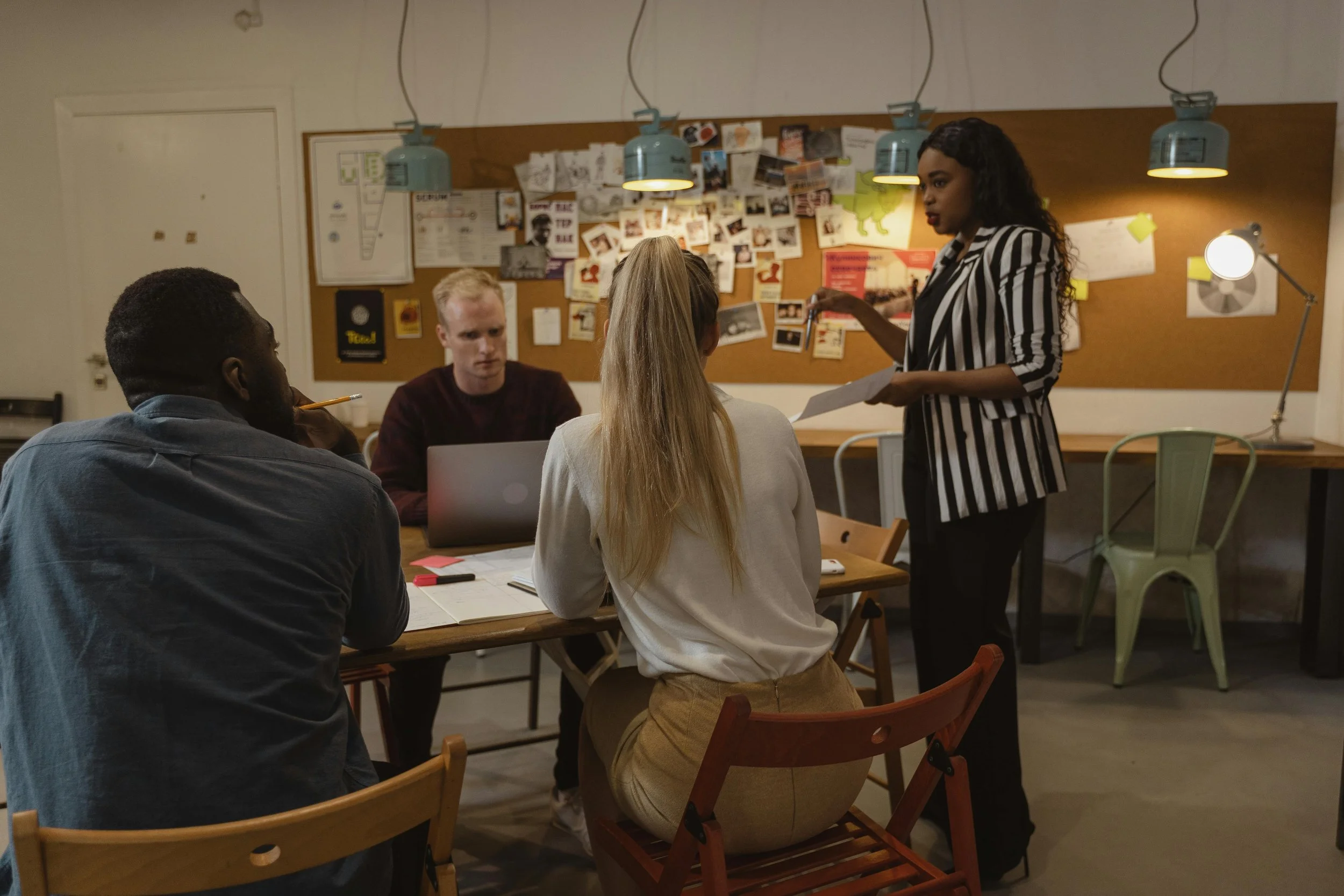 A diverse group of four young adults having a meeting in a casual office or co-working space, with one woman standing and speaking to three seated individuals, in front of a bulletin board decorated with photos and notes.