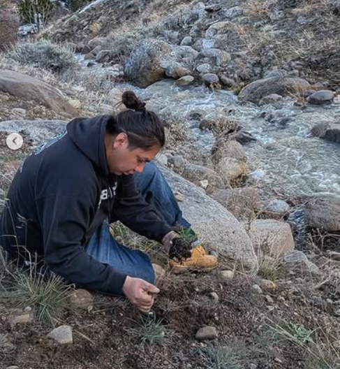 Person planting or tending to small plants near a rocky stream in a mountainous area.