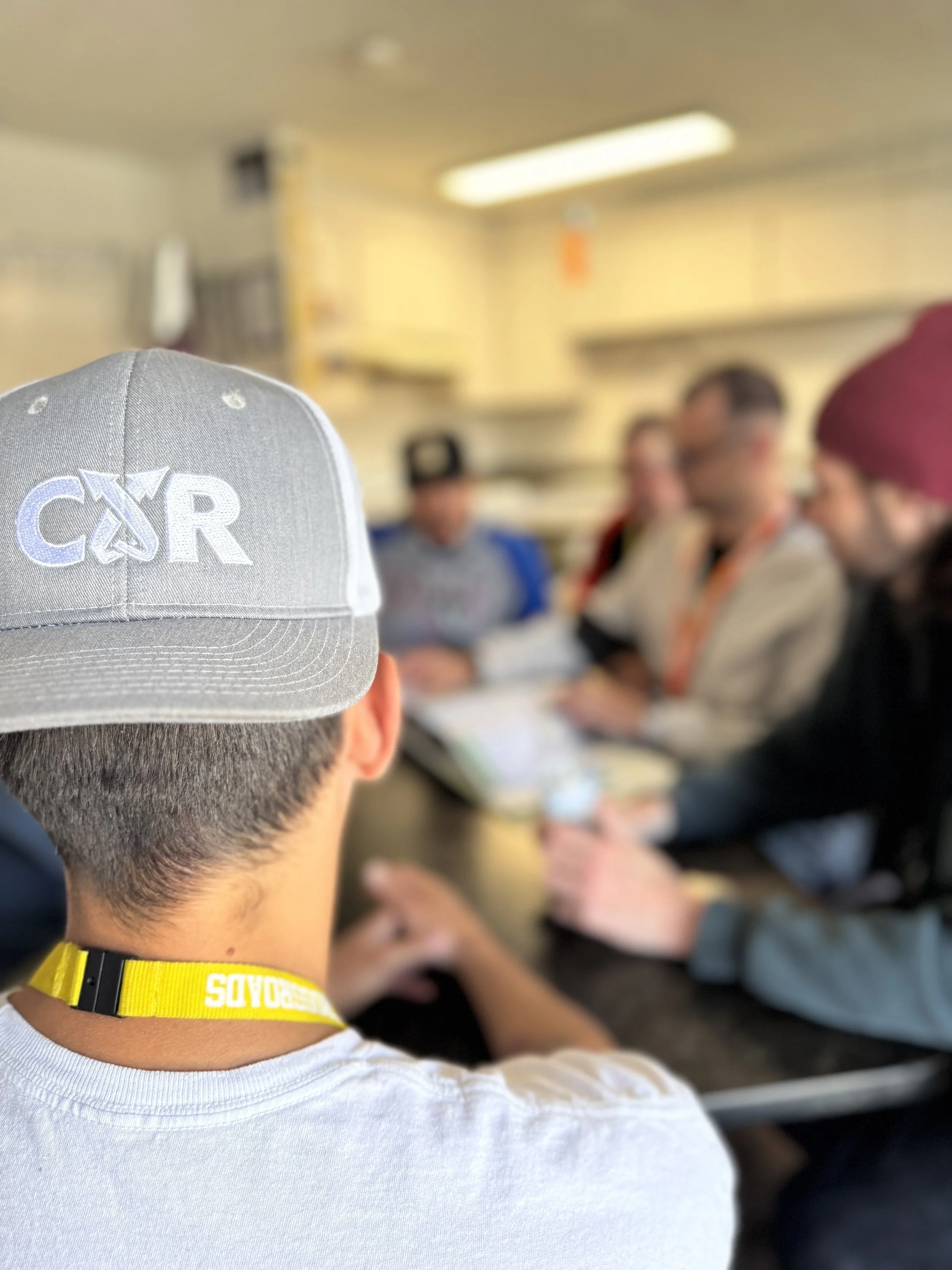 Person wearing a gray baseball cap with 'CXR' logo and a yellow lanyard, sitting at a table in a classroom or meeting room with others, blurred in the background.