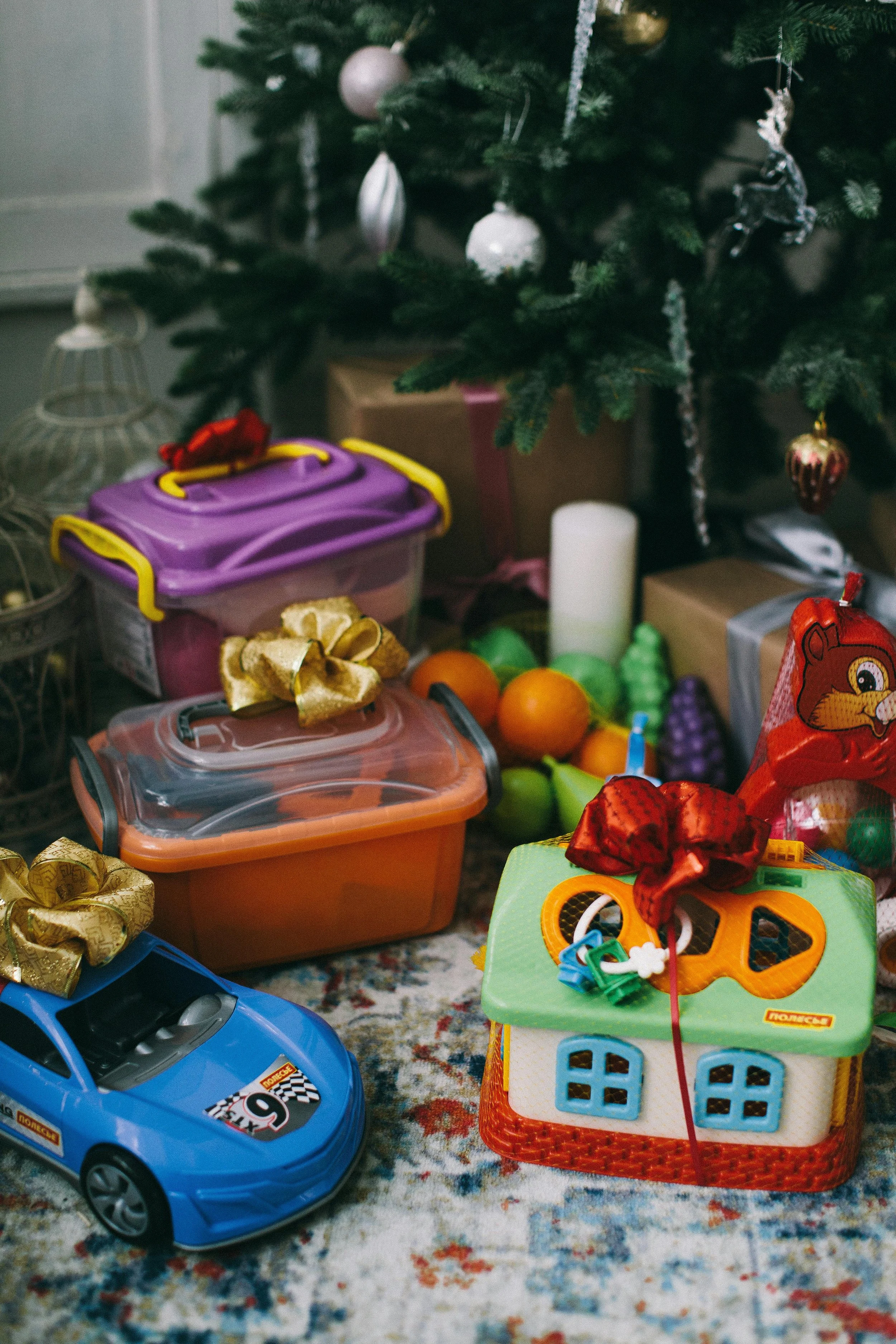 Children's toys placed under a decorated Christmas tree, including a toy car, a house-shaped toy, and storage containers with gold bows, with wrapped presents and a candle in the background.