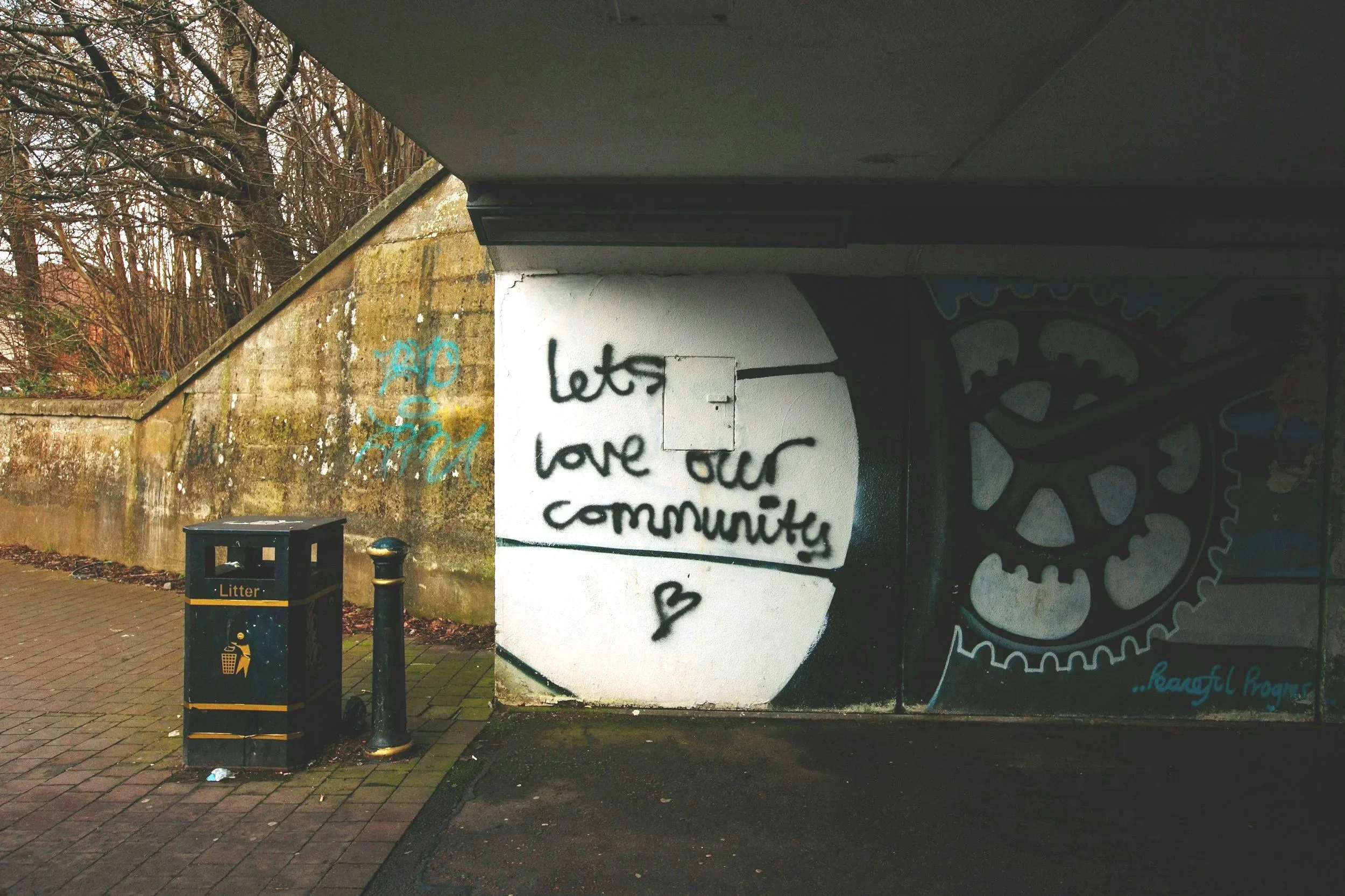 Graffiti on a wall under a bridge with a message that says, "lets love our community" and a small heart symbol. There is a trash bin labeled "Litter" and a black post nearby.