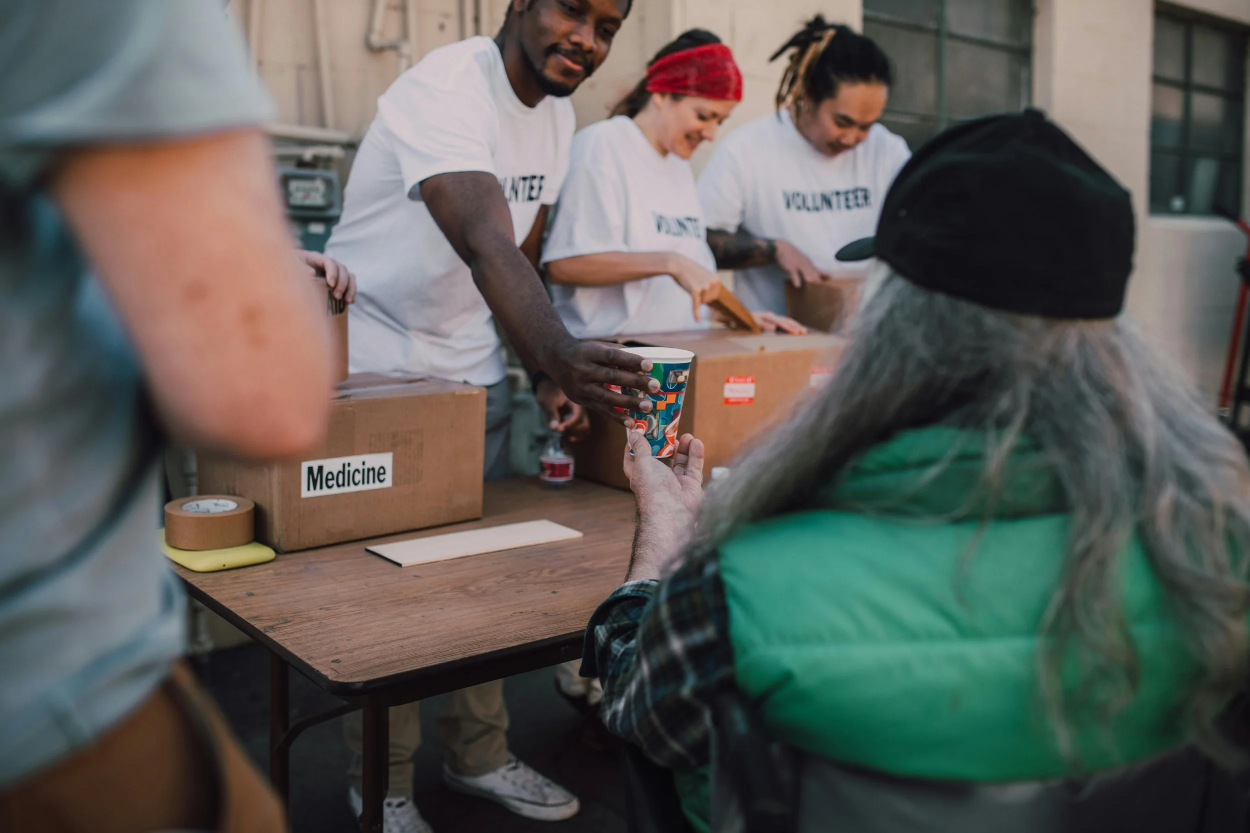 Volunteer workers distributing supplies to a person at a donation event, with boxes labeled 'Medicine' in the background.