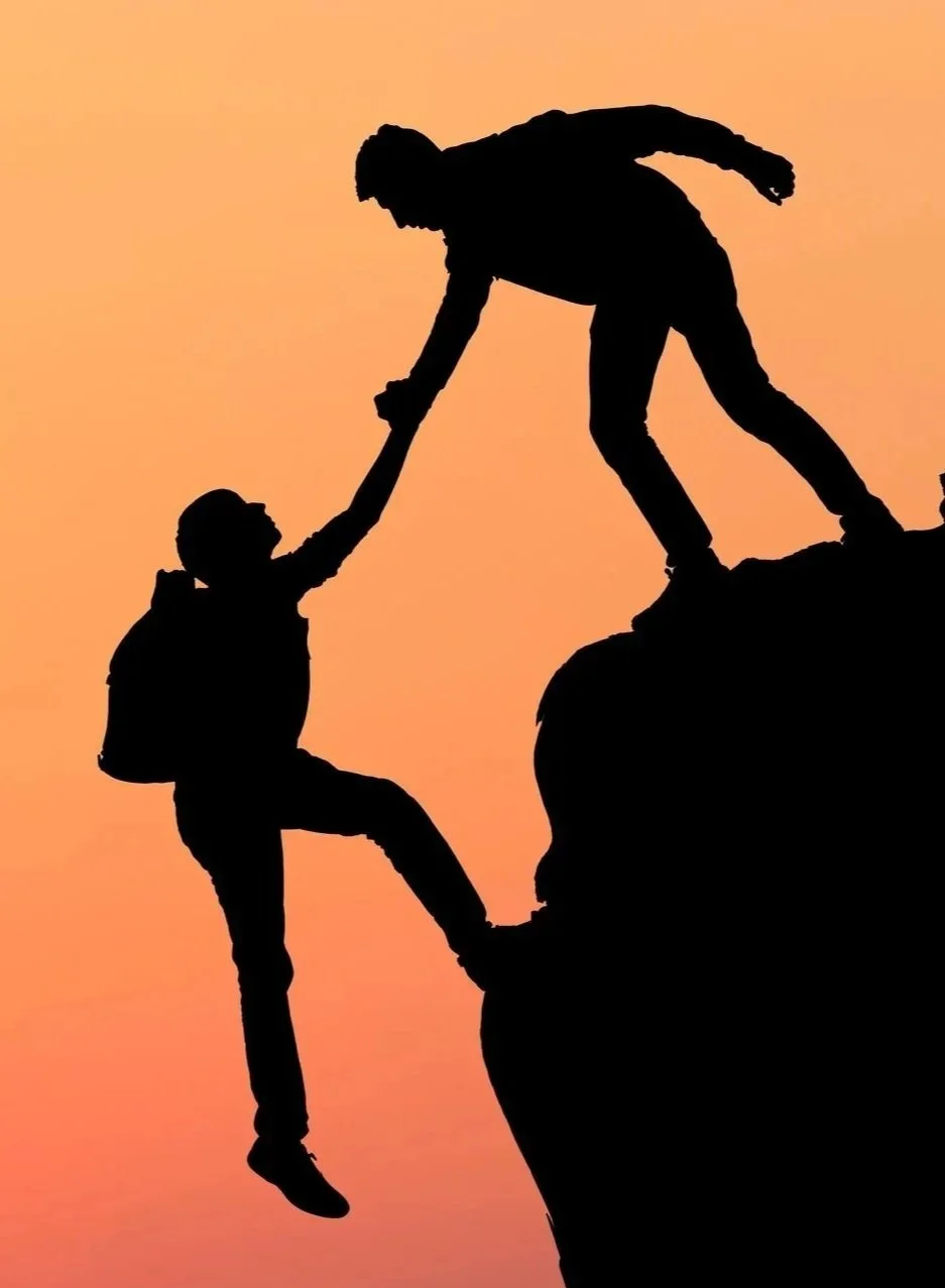 Silhouettes of two people helping each other climb a mountain at sunset.