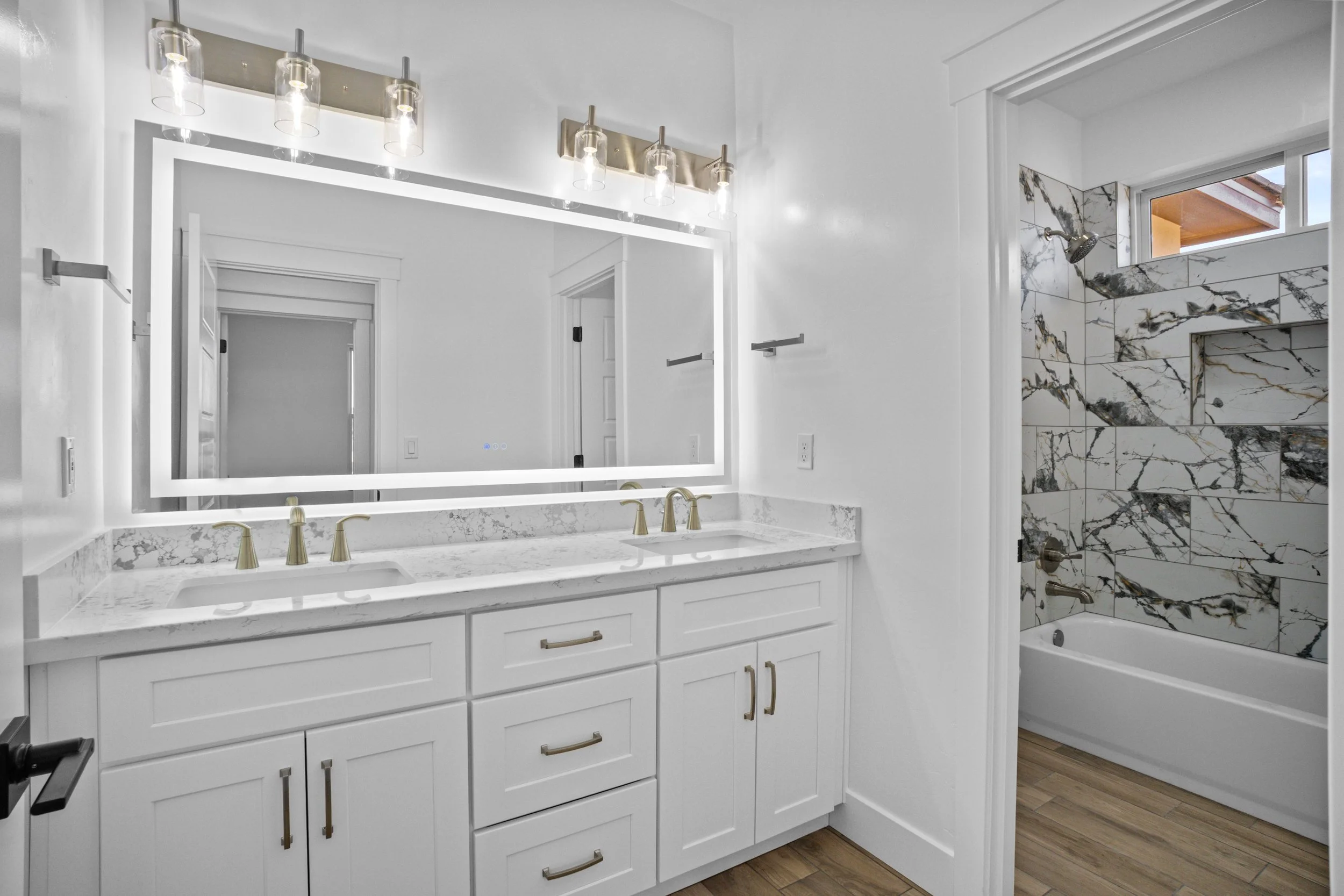 Modern bathroom with white cabinetry, marble countertop, large illuminated mirror, and a separate shower area with marble tiles and a bathtub.