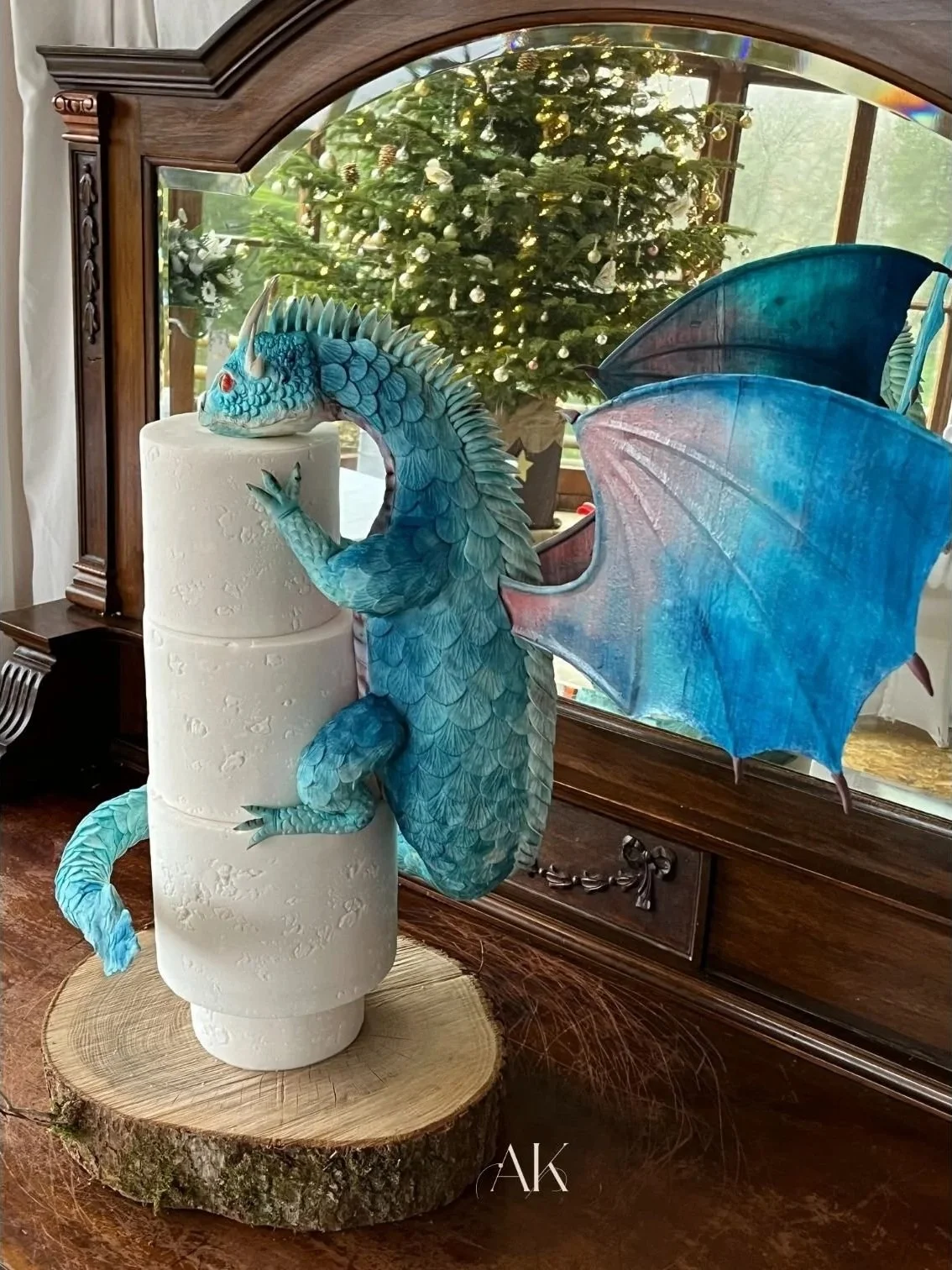 A decorative sculpture of a blue dragon with detailed scales, perched on a white textured cylindrical object, with large blue and purple wings spread out. The sculpture is placed on a wooden log base in front of a mirror reflecting a Christmas tree w
