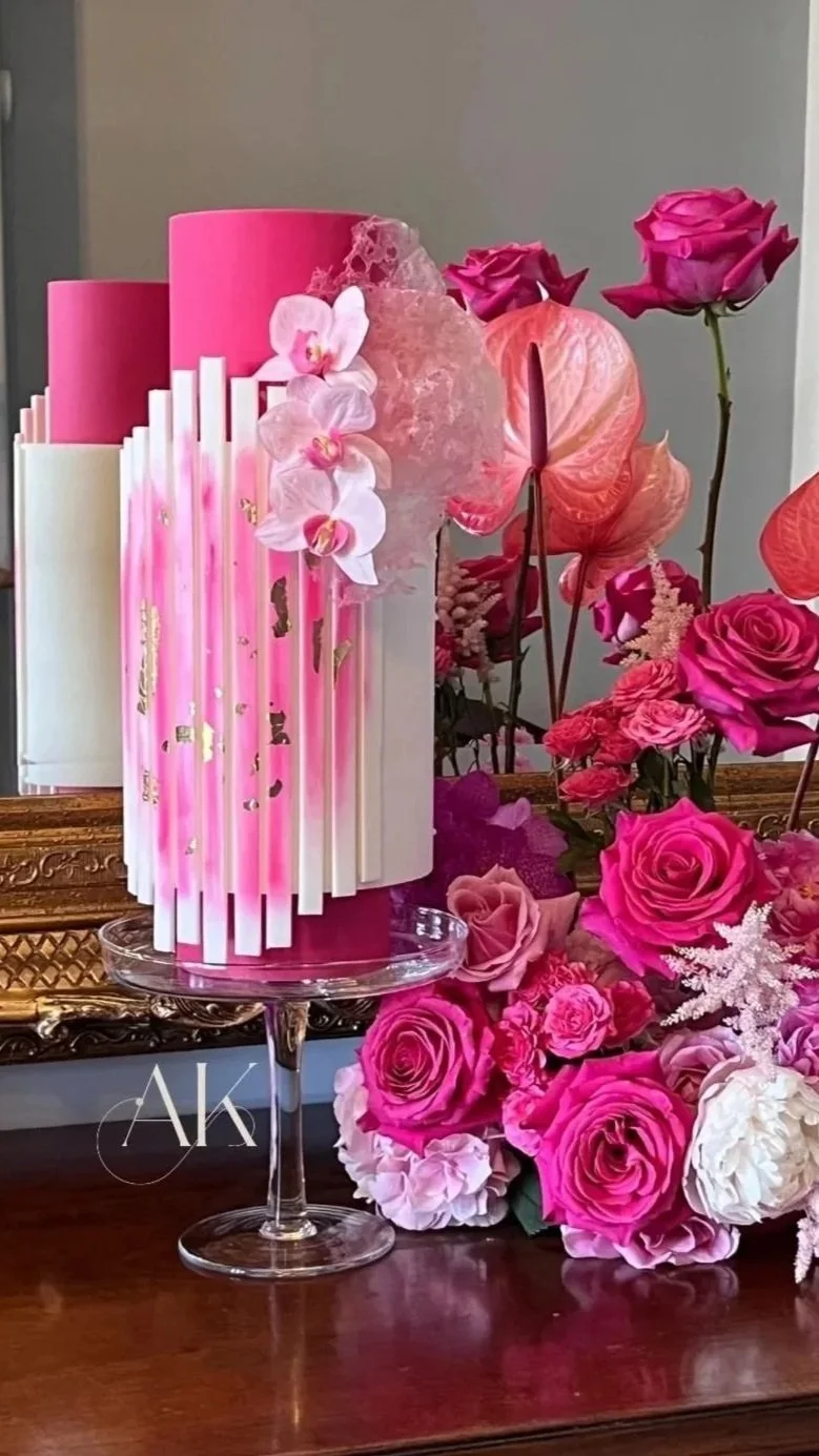 Elegant pink and white floral cake with orchids and roses, surrounded by pink roses and anthuriums on a wooden table.
