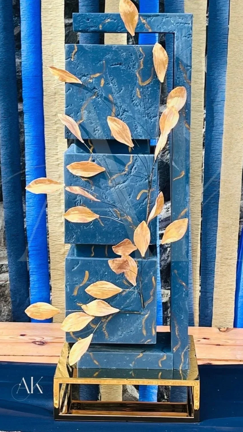 A decorative, sculptural cake with blue marble-like blocks and golden leaves on black stems, arranged vertically on a gold base. Background features vertically striped blue, black, and beige elements.