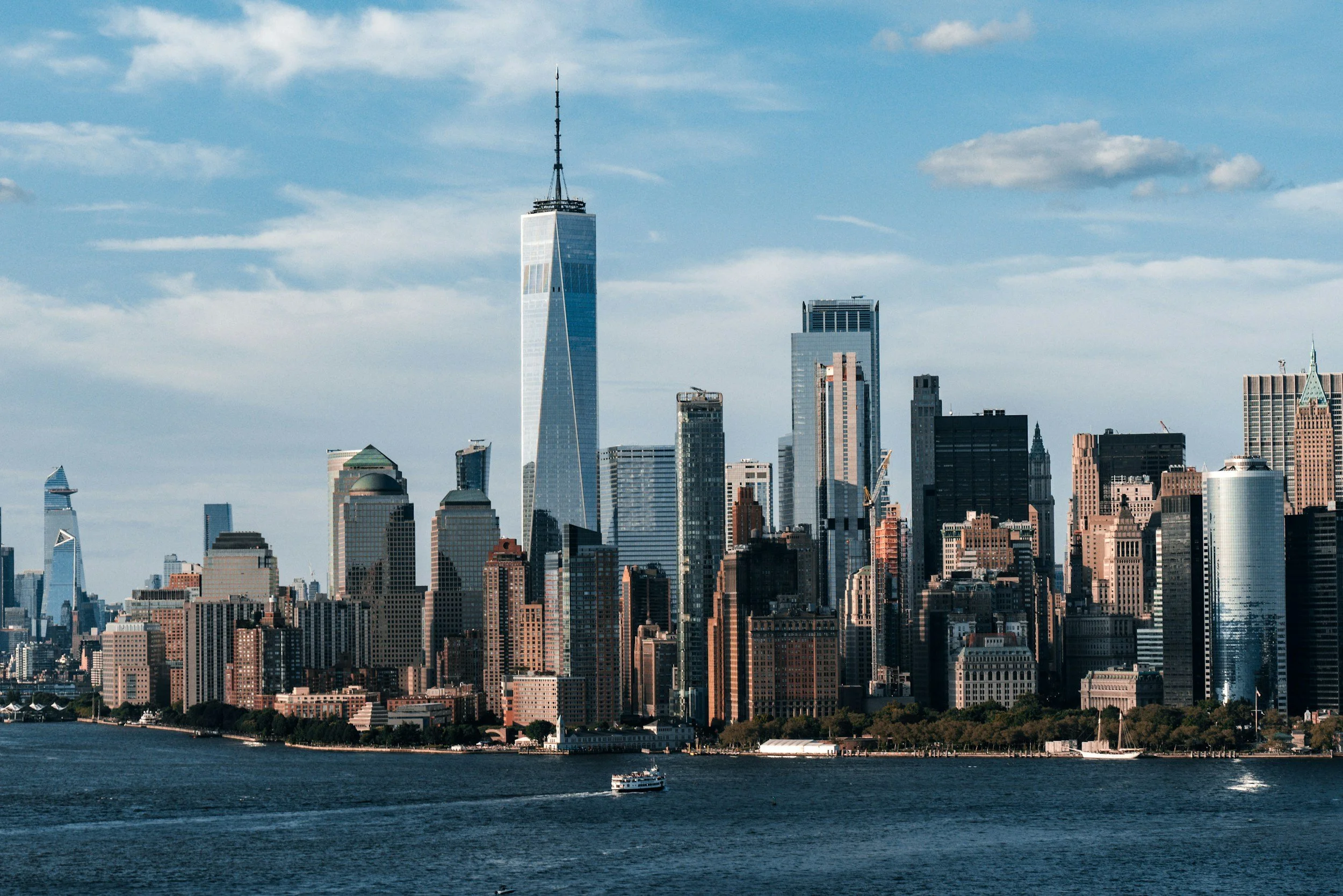 Skyscrapers of Manhattan skyline with One World Trade Center in the foreground and water with boat in the river