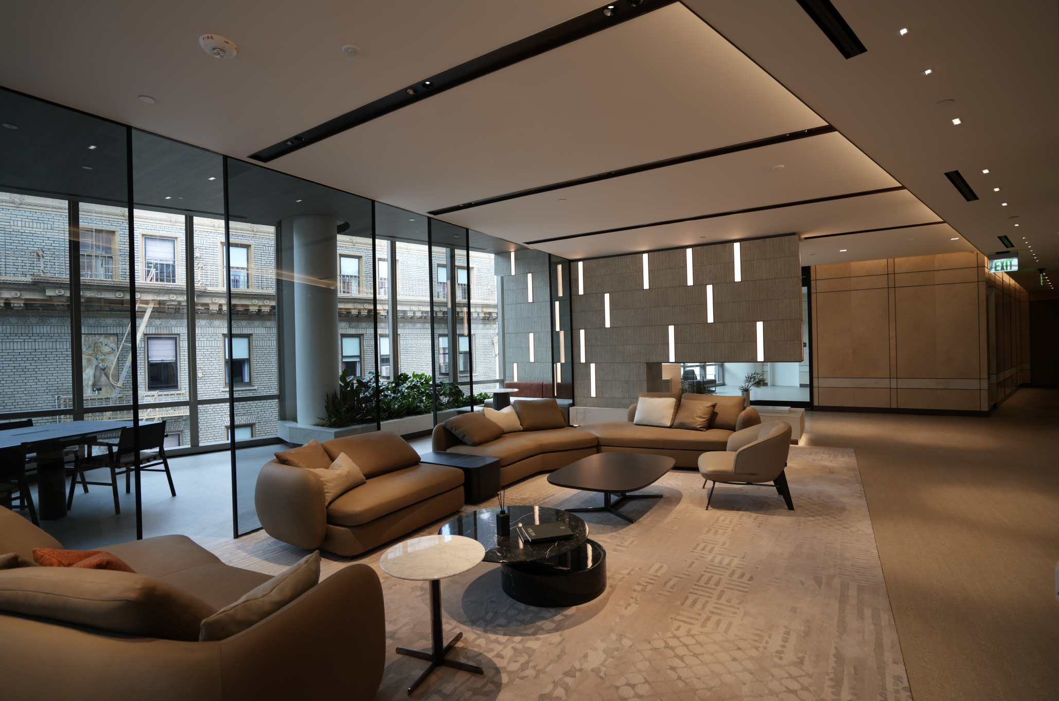 Modern lobby with beige sofas, a black coffee table, side tables, and large floor-to-ceiling windows, with city buildings outside and indoor plants.