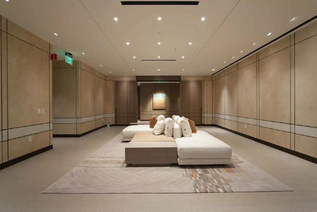 Beige walls, recessed lighting, and a large white sectional sofa with throw pillows, situated on a neutral-toned area rug.